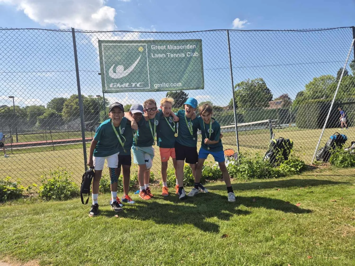 Bucks County Tennis players at a tennis club in Great Missenden wearing medals, standing on grass at a tennis court, smiling with arms around each other, celebrating their win.