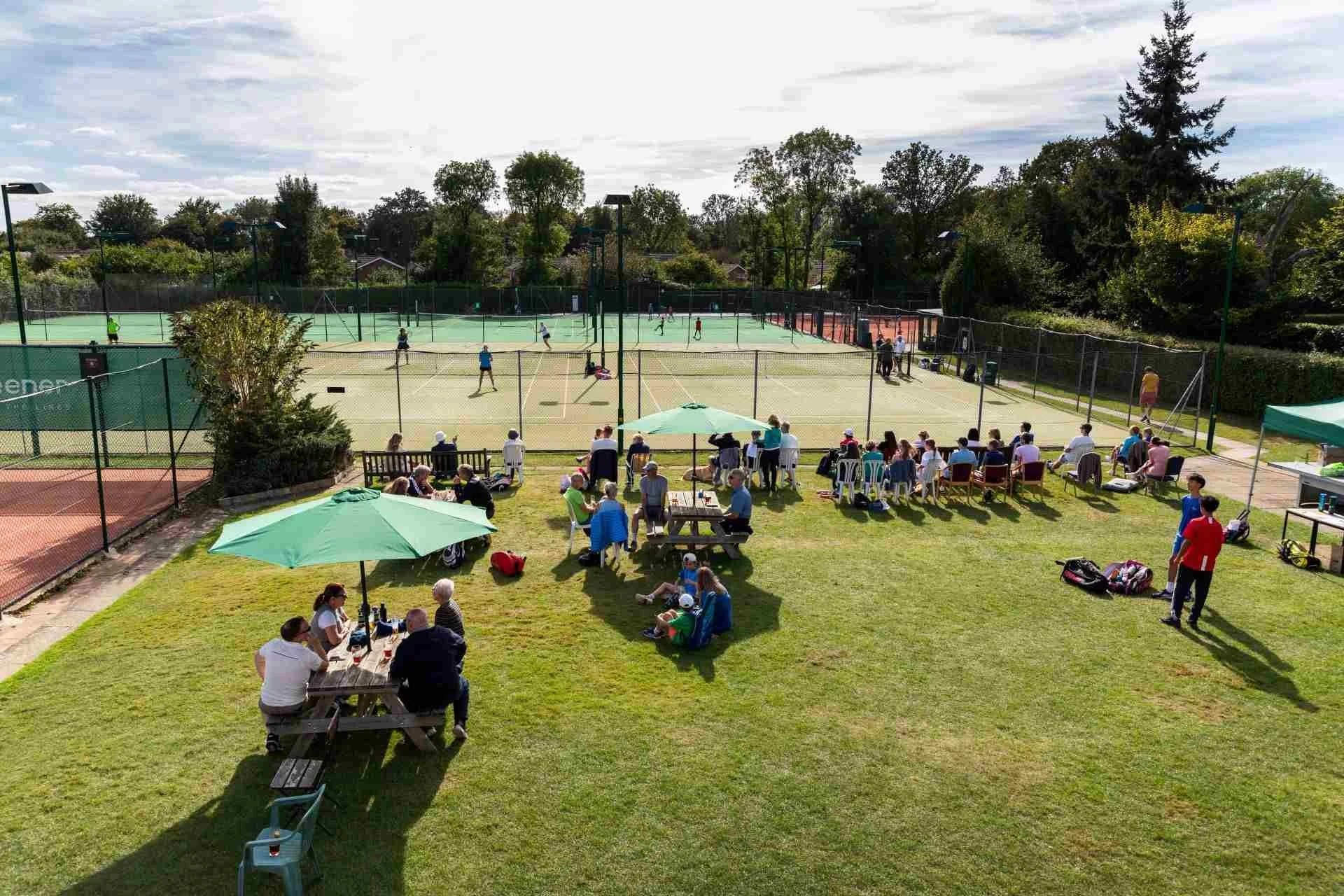 People playing tennis on outdoor courts, with spectators and others sitting under umbrellas on grass in the foreground, surrounded by trees under a partly cloudy sky.