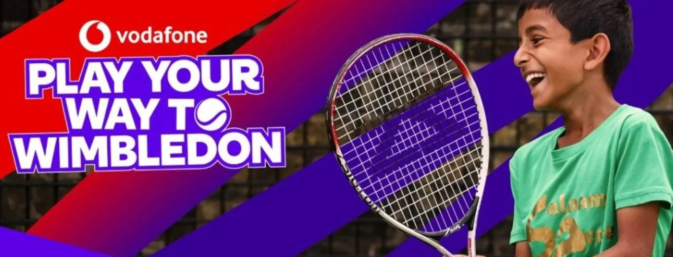 A young boy tennis player laughing with a tennis racket in front of a background with bold red and purple graphics and text that reads: 'Vodafone, Play Your Way to Wimbledon'.