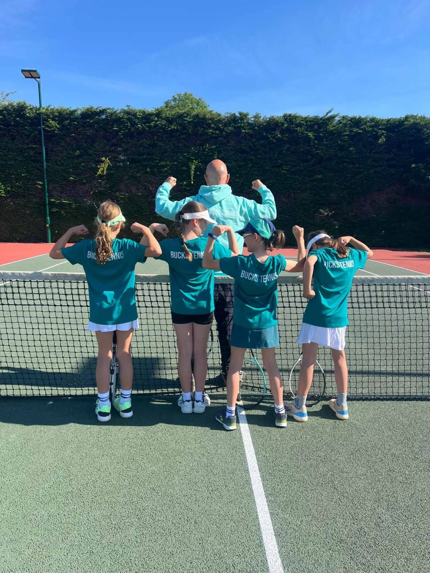 Bucks County Players and Coach tennis court with their backs to the camera, flexing their biceps. They are wearing teal shirts with 'BUCKSTENNIS' written on them and are standing behind a tennis net