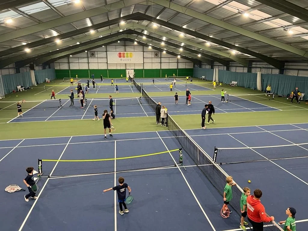 Bucks Indoor tennis facility with multiple courts, children and adults playing tennis, some practicing, and others coaching, under a high arched roof with bright lighting in High Wycombe