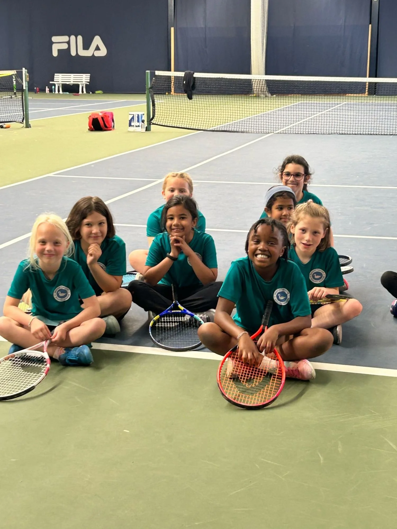 A group of seven under 8 bucks county tennis girls sitting on an indoor tennis court with tennis rackets. They are smiling and wearing matching teal shirts and black shorts, and are positioned in front of black tennis nets.