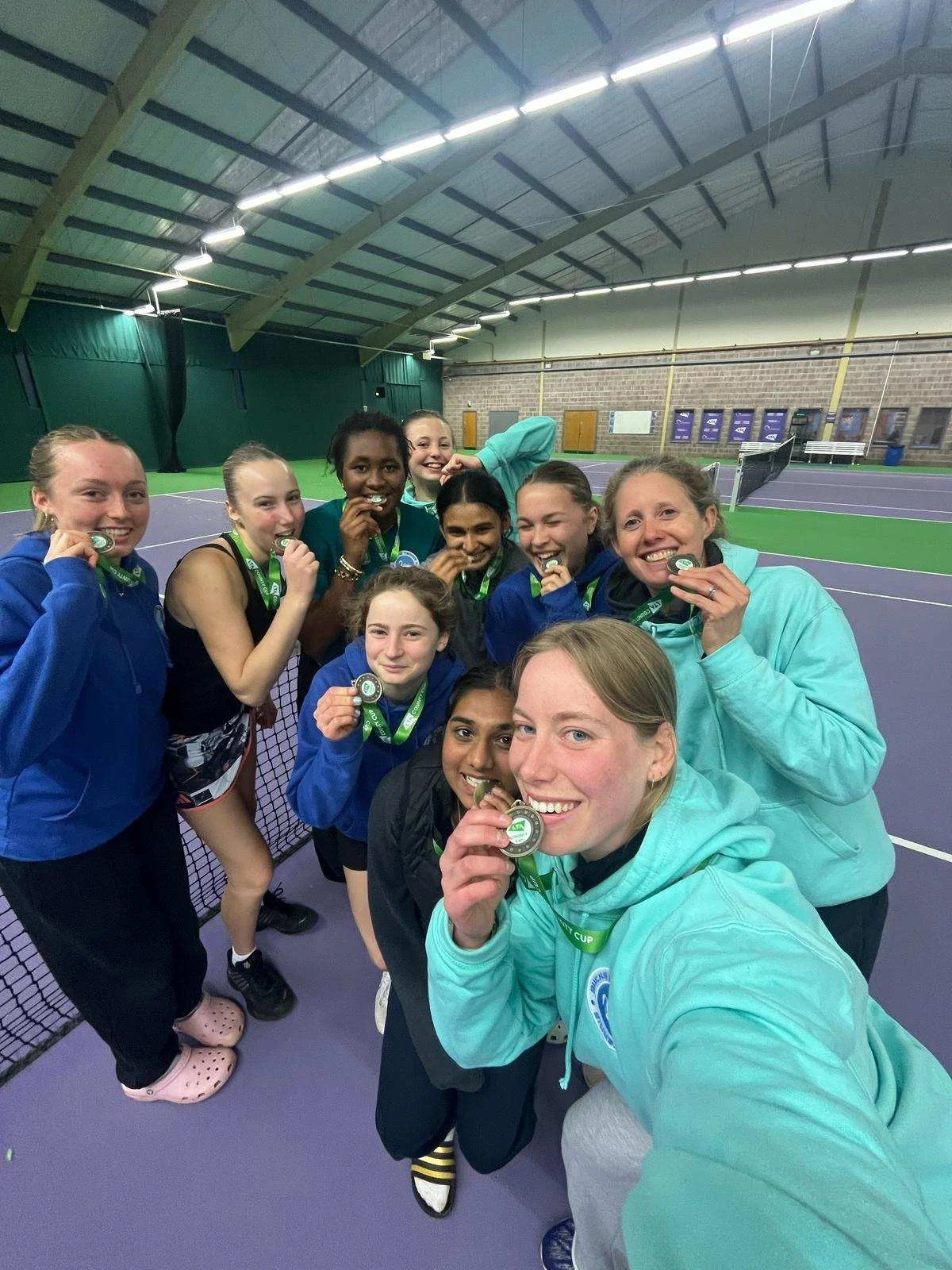 Bucks County Tennis Players with medals around their necks, celebrating on an indoor tennis court. They are smiling, biting their medals, and squatting or standing close together.