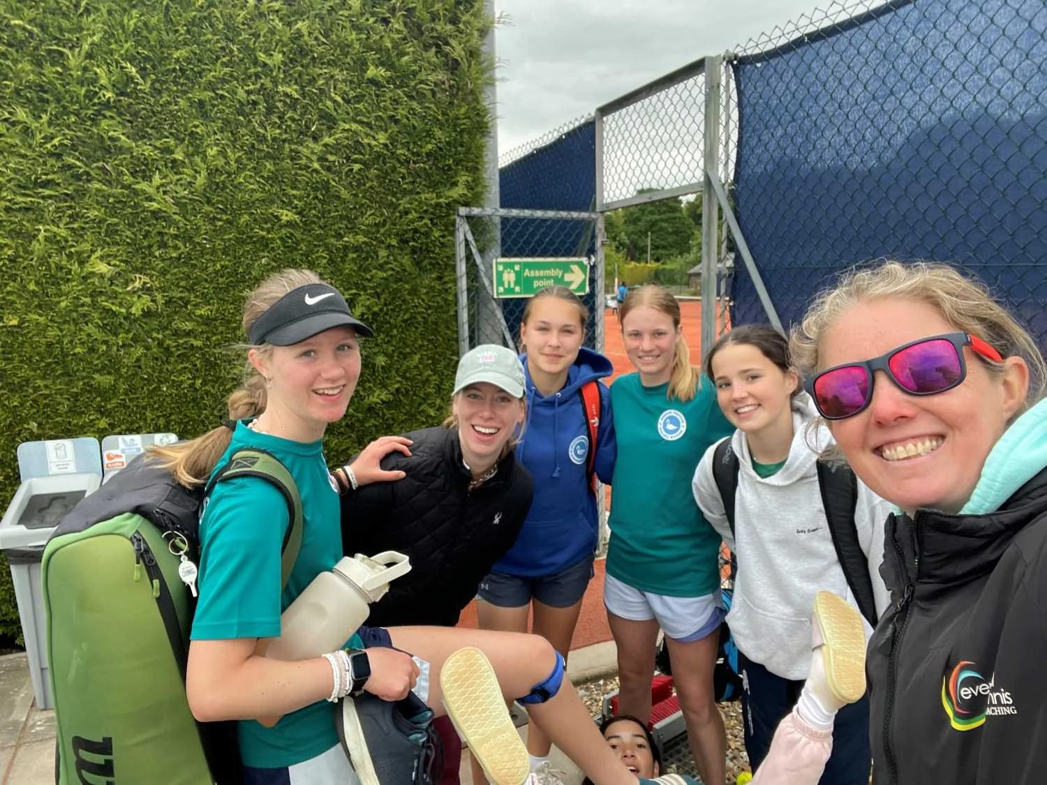 Group of six Bucks County Tennis players women smiling for a group photo at a tennis court, with greenery and a fence in the background.