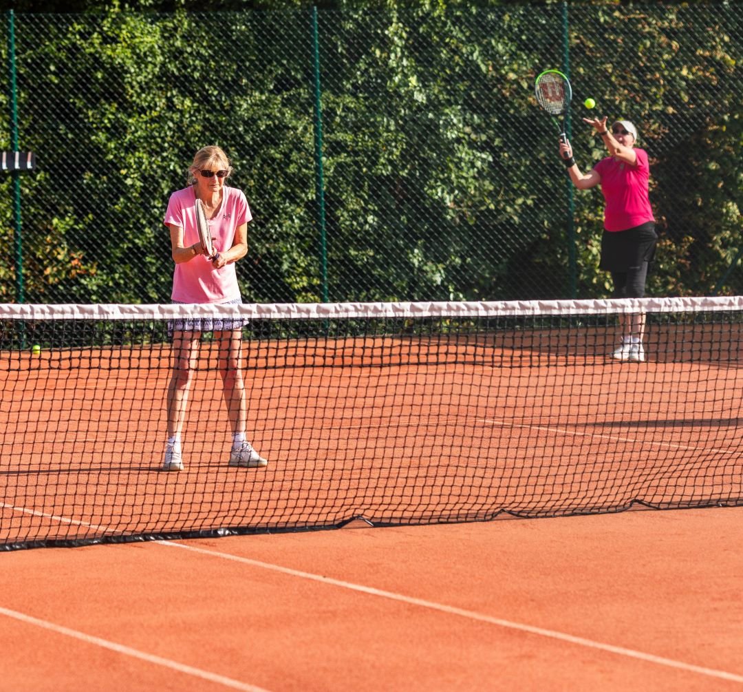 Two women playing tennis on an outdoor clay court surrounded by trees, one woman preparing to return a shot, and the other hitting the ball with a racket.