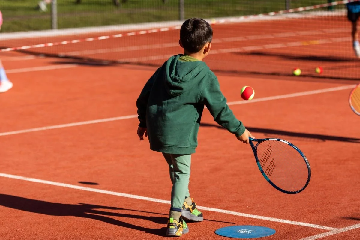 A young boy on a red clay tennis court at Chesham Bois Tennis Club in Bucks, holding a tennis racket, with tennis balls around him, wearing a green hoodie and gray pants.