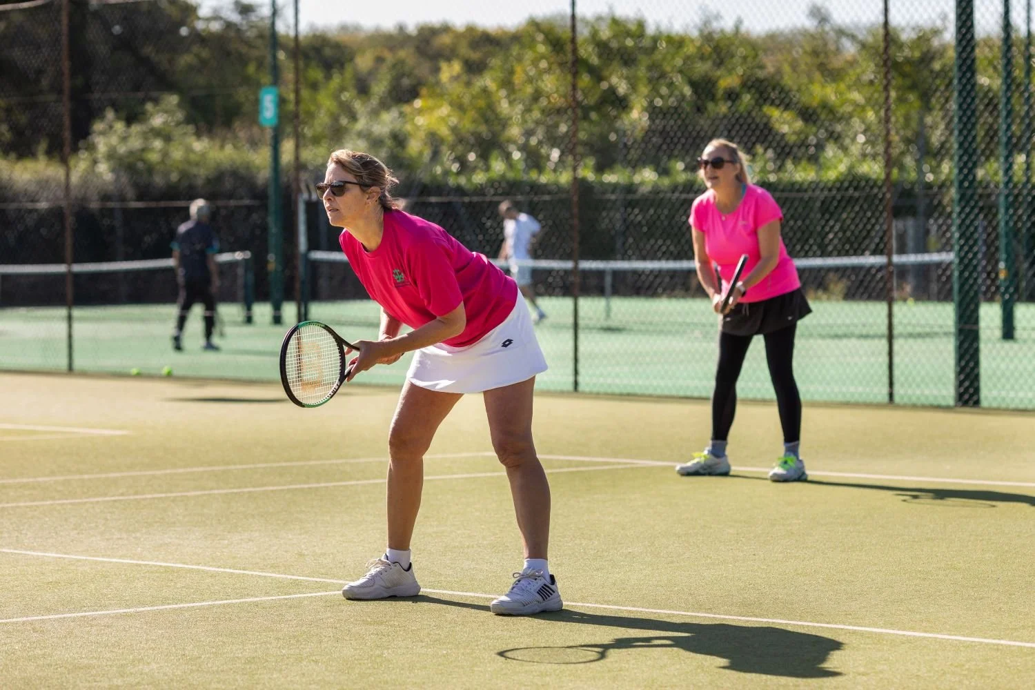 Two women playing pickleball on an outdoor court, both wearing sunglasses and pink shirts. One woman is preparing to hit the ball while the other stands behind her, holding a paddle.