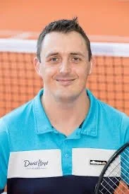 A man standing on an indoor clay tennis court holding a tennis ball and a racket, with a basket of tennis balls nearby.