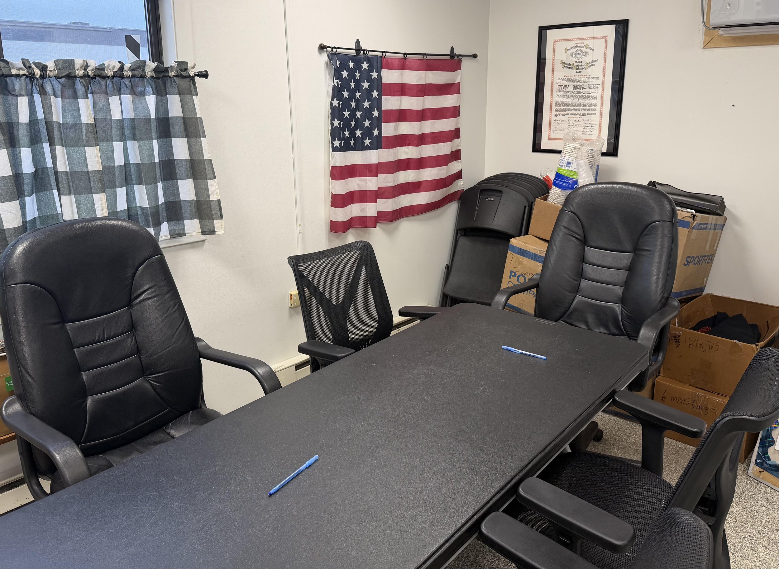 An office for local 2403, meeting room with black chairs around a dark table, a window with checkered curtains, an American flag and a partially visible map or certificate on the wall, and stacked boxes and reusable water bottles in the corner.