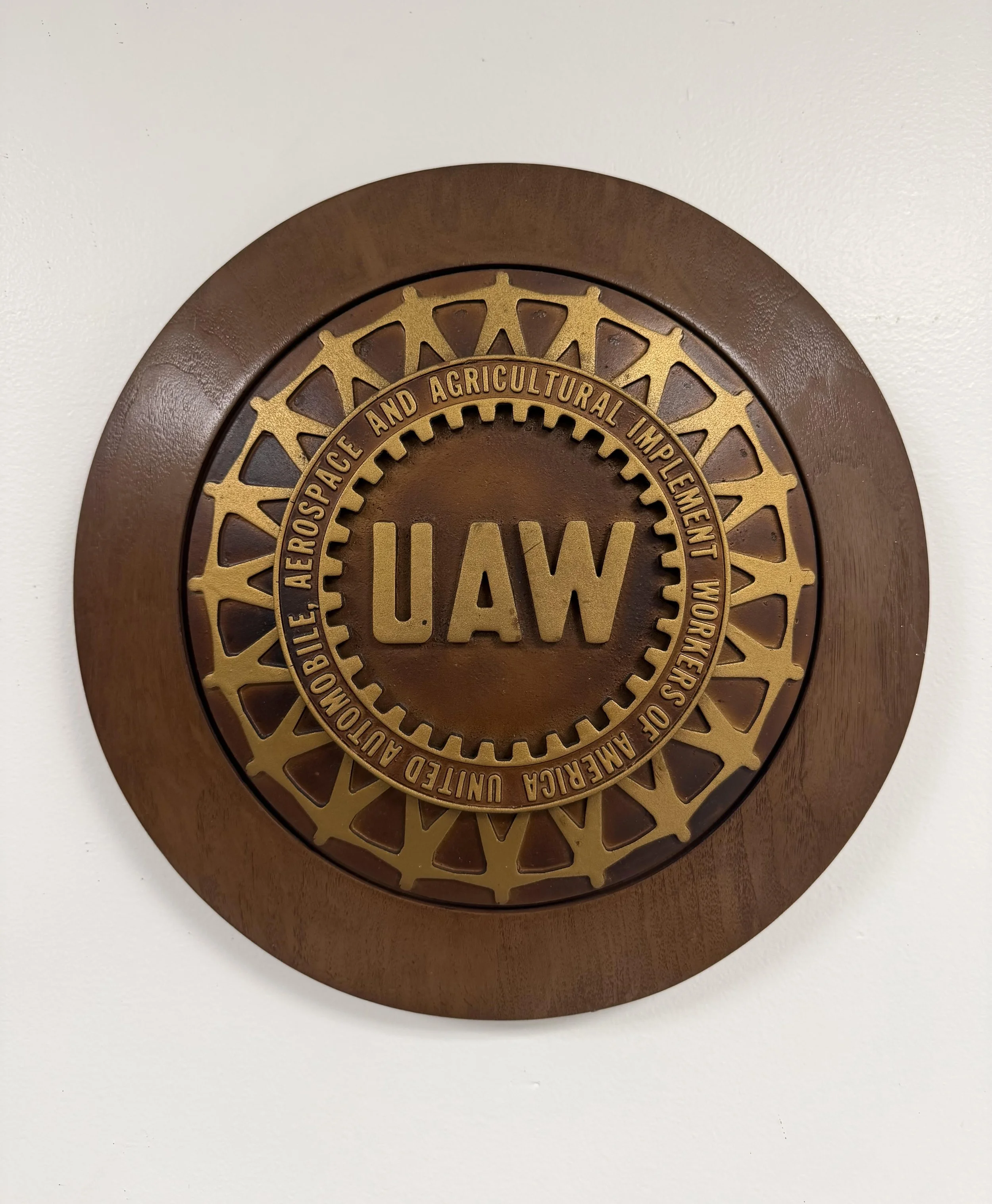 Close-up of a circular wooden plaque or sign with a gold and black emblem for UAW, United Auto Workers, featuring a gear and the text, "UAW".