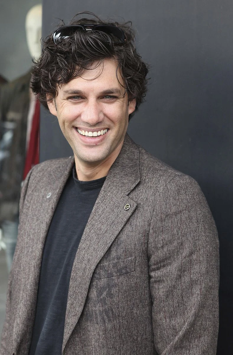 A smiling man with curly brown hair wearing sunglasses on his head, a brown blazer, and a black shirt, standing against a dark background.