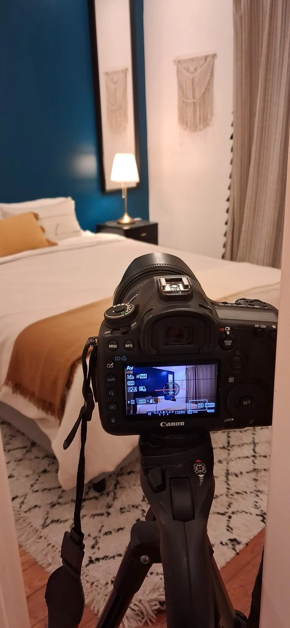A camera on a tripod capturing an image of a neatly made bed with a beige blanket, in a bedroom with a blue accent wall, a black nightstand, a white table lamp, and framed wall art.