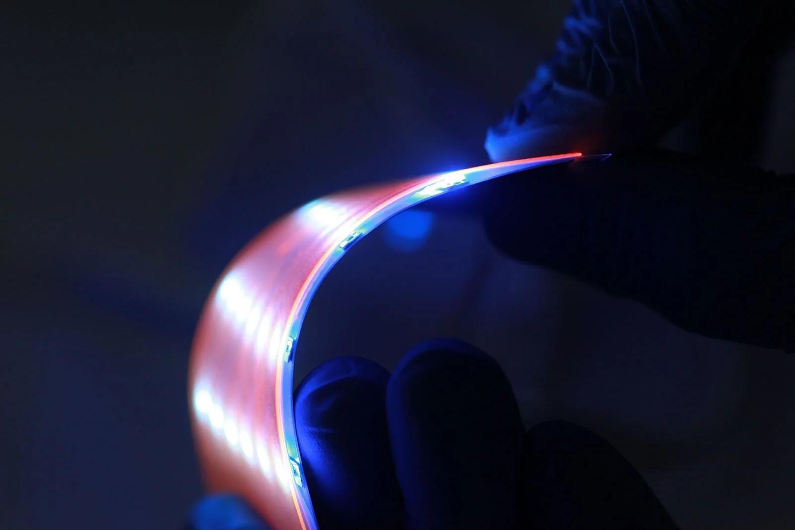 Close-up of a person's fingers holding a flexible, illuminated electronic circuit with colorful LED lights in a dark environment.