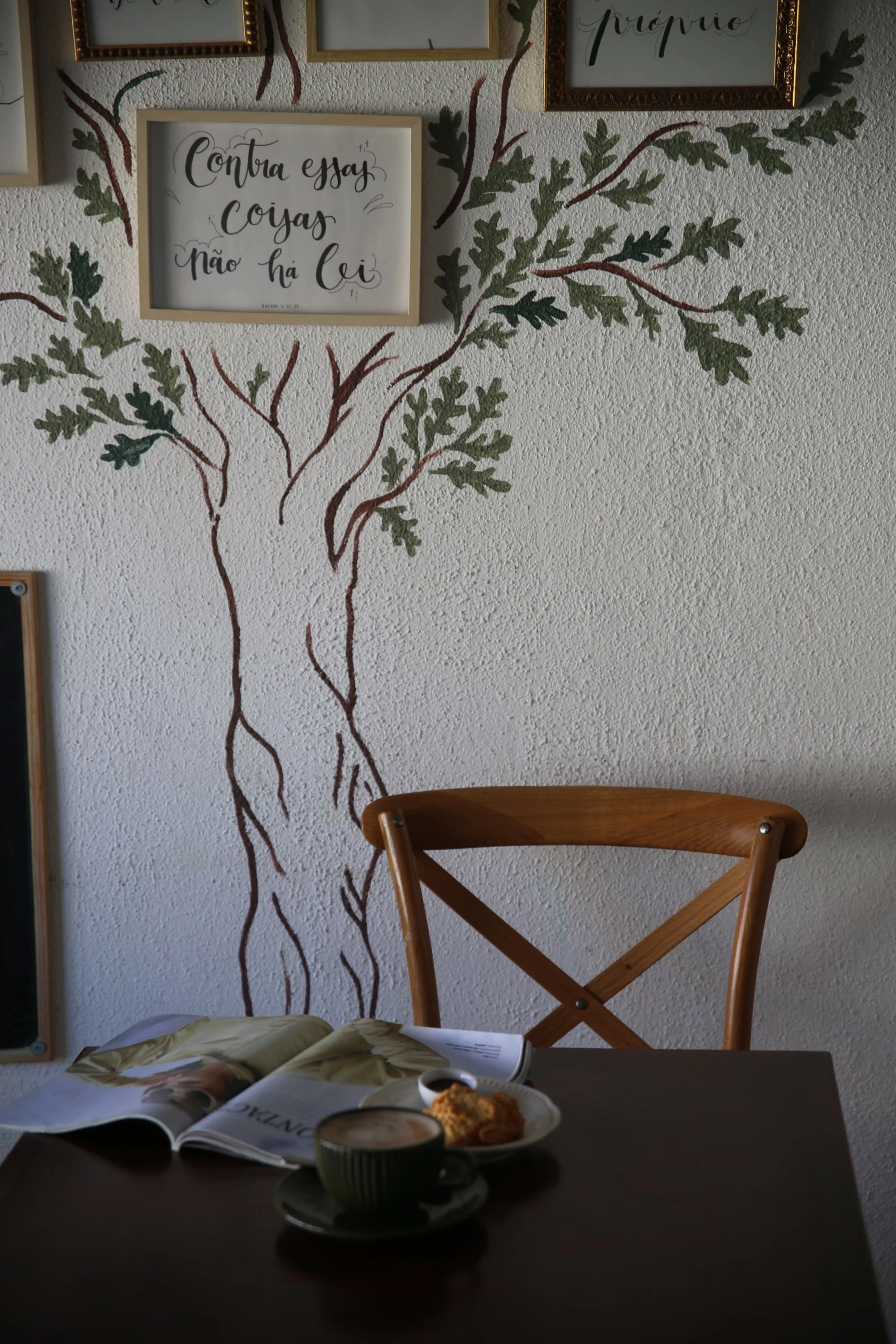 A table with an open magazine, a cup of coffee, and a plate with food. Behind the table, there's a wall with a painted tree and framed wall art with handwritten text.