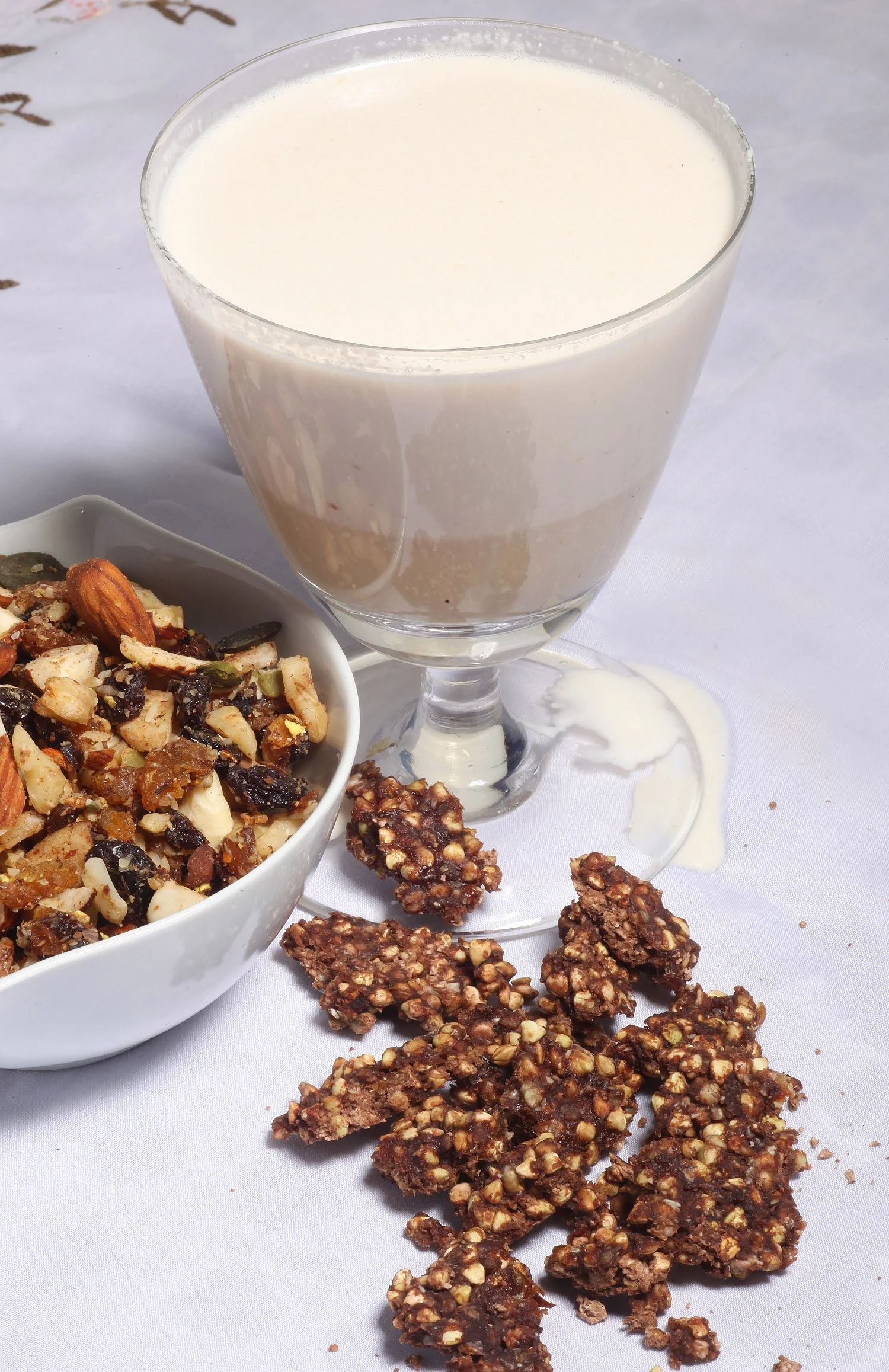 A glass of milk, a white bowl filled with a nut and dried fruit trail mix, and clusters of peanut and oat granola bars scattered on a white tablecloth.
