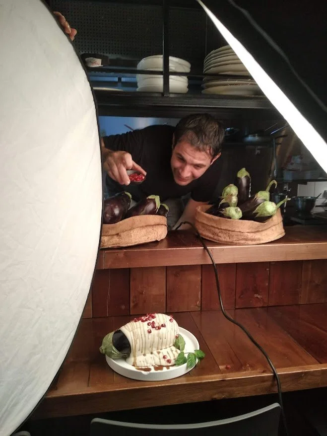 A man peering from behind a counter at a display of eggplants and a decorated cake resembling an eggplant. The scene is set in a kitchen with shelves and dishes in the background.
