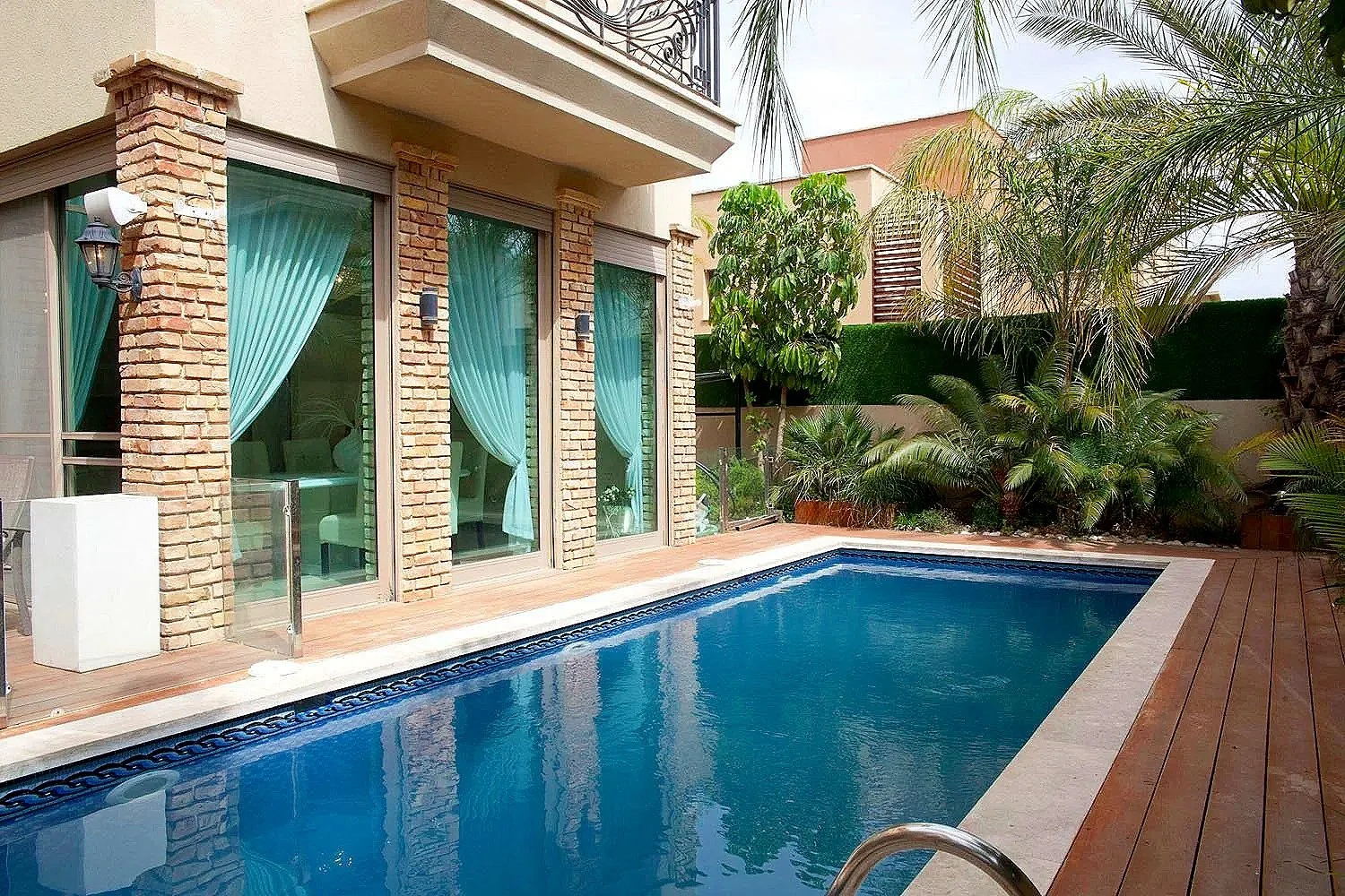 A backyard with a swimming pool surrounded by a wooden deck, green plants, palm trees, and a house with brick and glass windows with blue curtains.