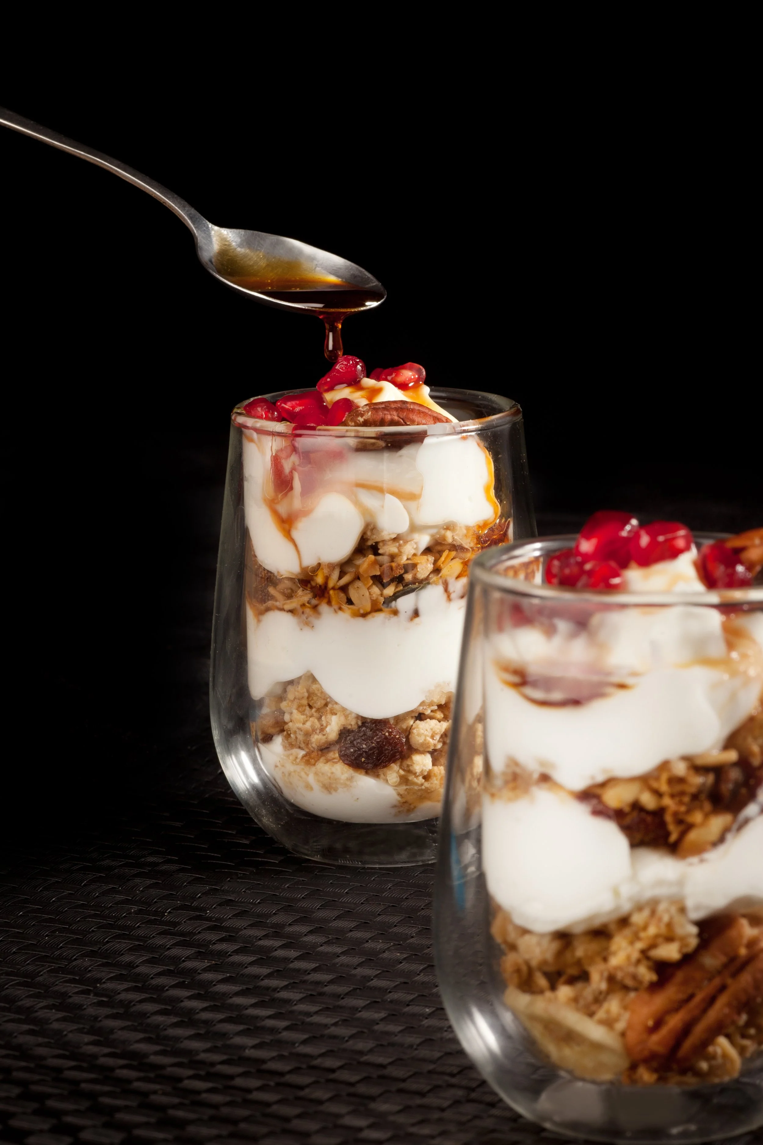 Close-up view of dessert parfaits in glass cups, showing layers of granola, yogurt, and fruit, topped with pomegranate seeds and caramel drizzle against a black background.