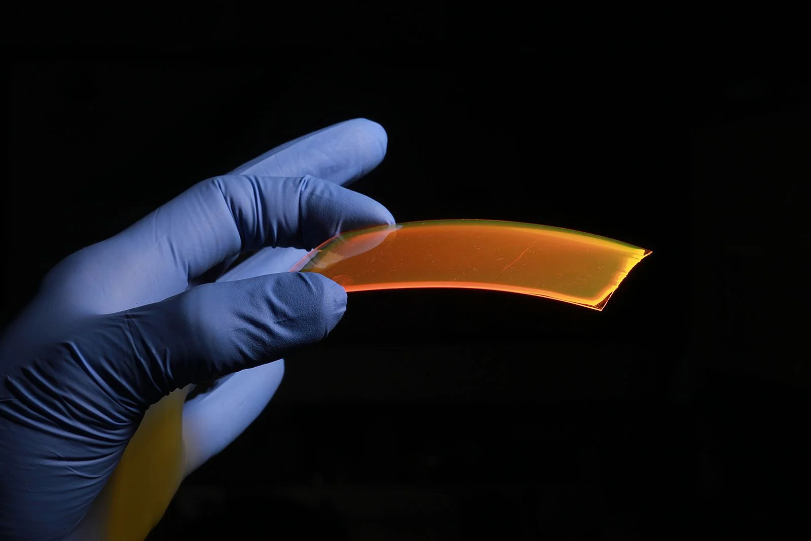 A gloved hand holding a curved, transparent piece of flexible electronic display or sensor, glowing with orange and green light against a dark background.