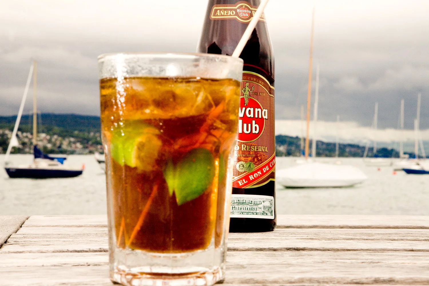 A glass of Rum and Coke with lime and a straw on a wooden table in front of a bottle of Havana club Rum, with boats and water in the background under cloudy skies.