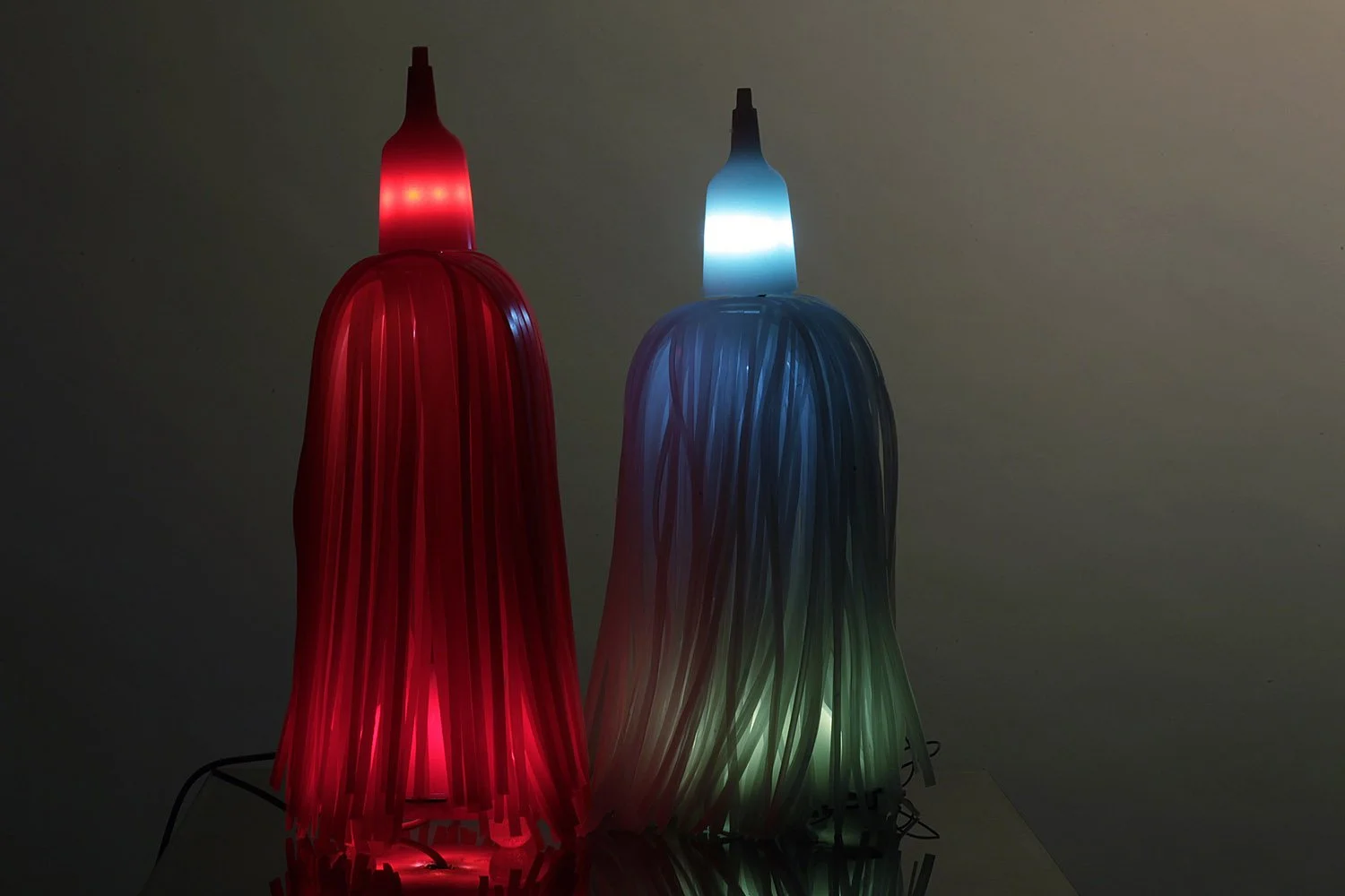 Two colorful table lamps with elongated, lampshade-like designs glowing in red and blue-green light in a dark room.