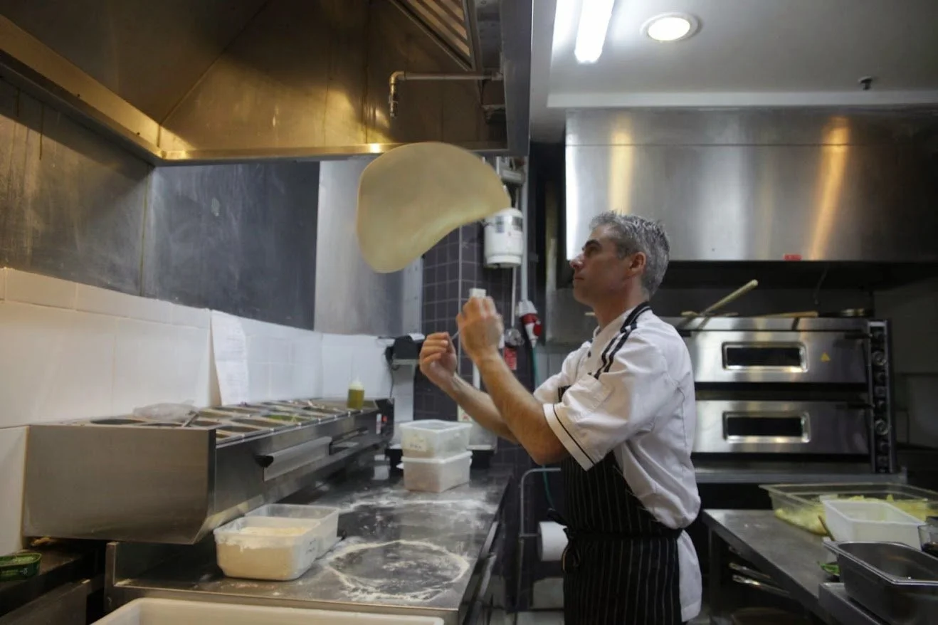 A chef in a commercial kitchen tossing pizza dough into the air.