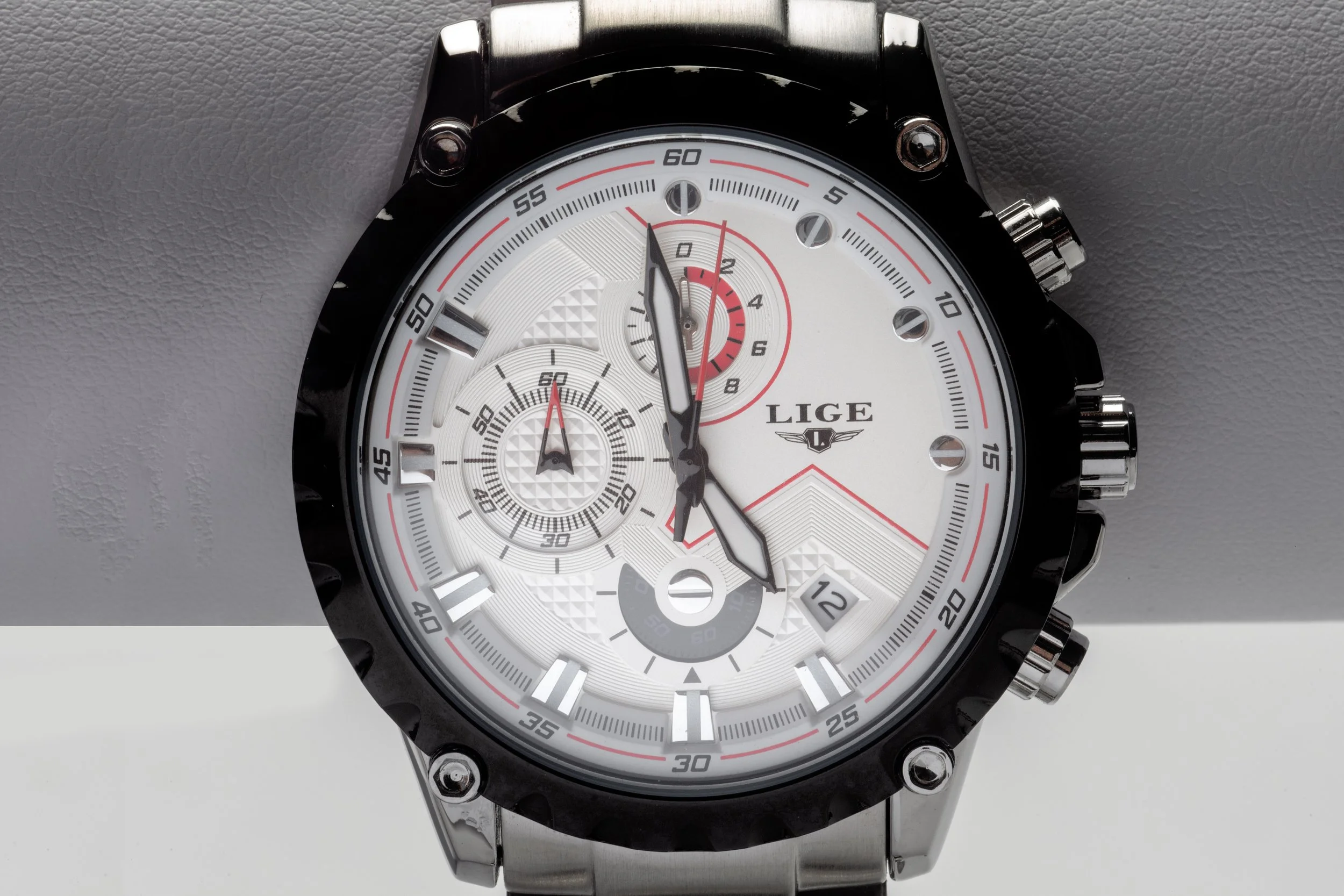 A men's analog wristwatch with a white dial, black bezel, and stainless steel bracelet. The dial has multiple subdials, red accents, and a date window, with the brand name "LIGE".