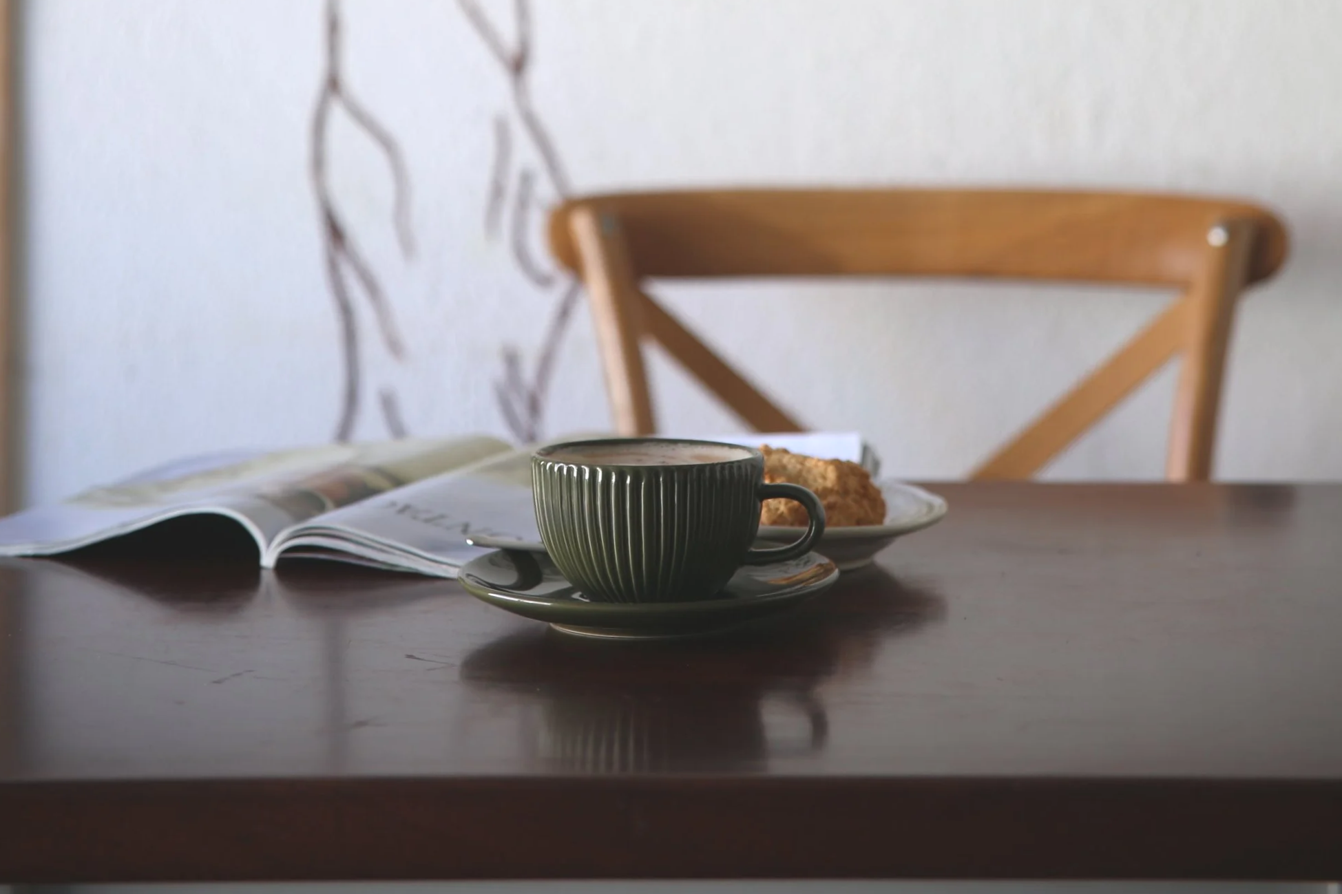 A table setting with a dark green cup of coffee on a matching saucer, an open magazine, and a plate with a slice of cake in the background. The background includes a wooden chair and a white wall with a decorative branch.