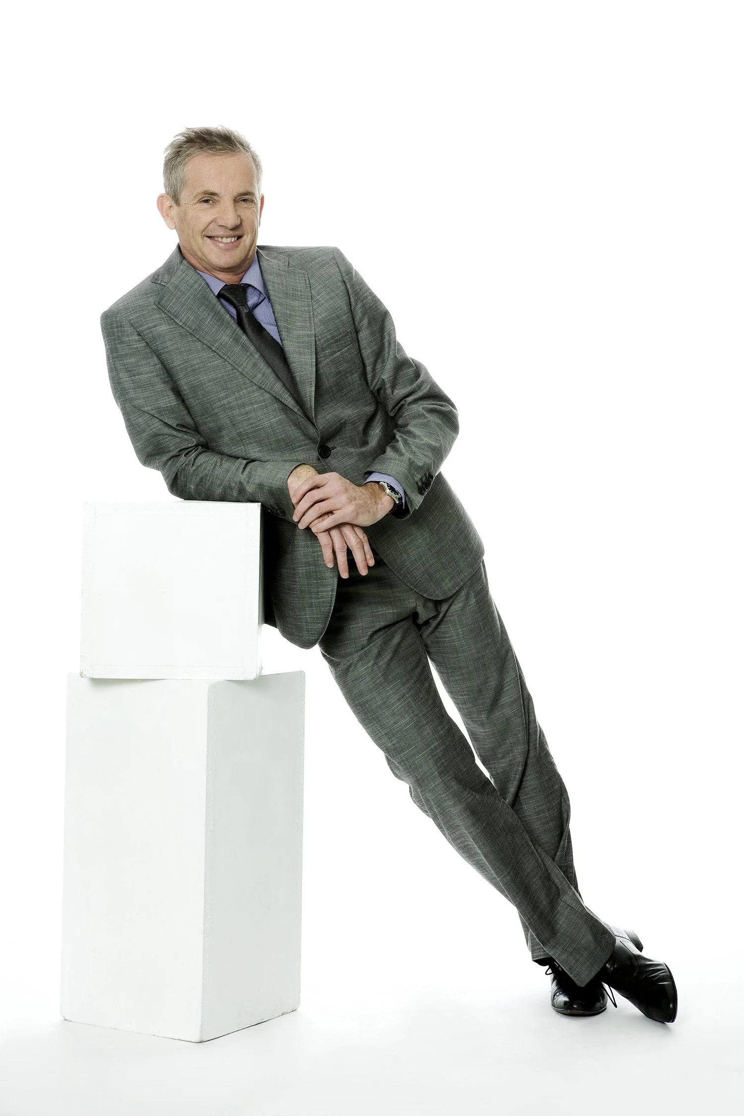 A middle-aged man in a gray plaid suit with a blue shirt and black tie, smiling while leaning on three white rectangular blocks against a white background.