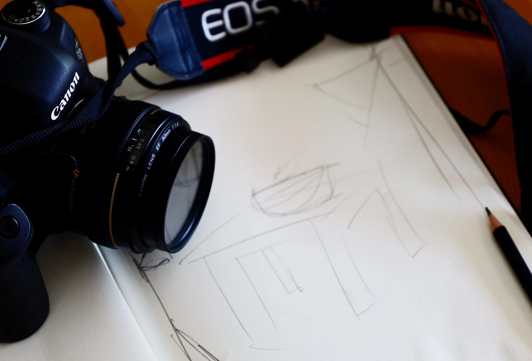 A Canon camera with a 50mm lens, a blue EOS strap, a sketchbook with a pencil drawing of geometric shapes, and a black pen on a white surface.