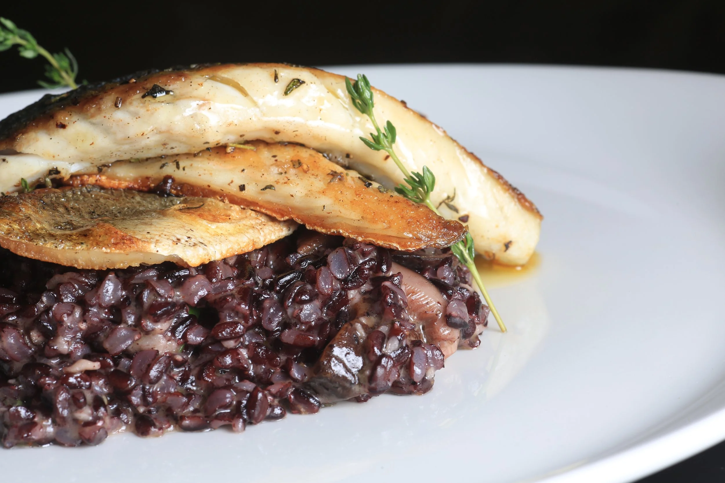 Crispy pan-seared white fish fillets resting on a bed of black rice, finished with a sprig of fresh thyme.