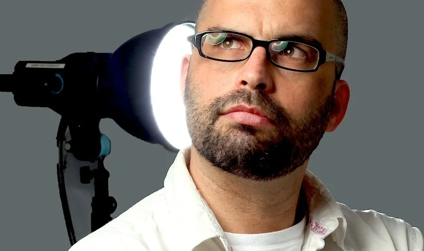 A man with glasses looking to the side with a photography or studio light in the background.
