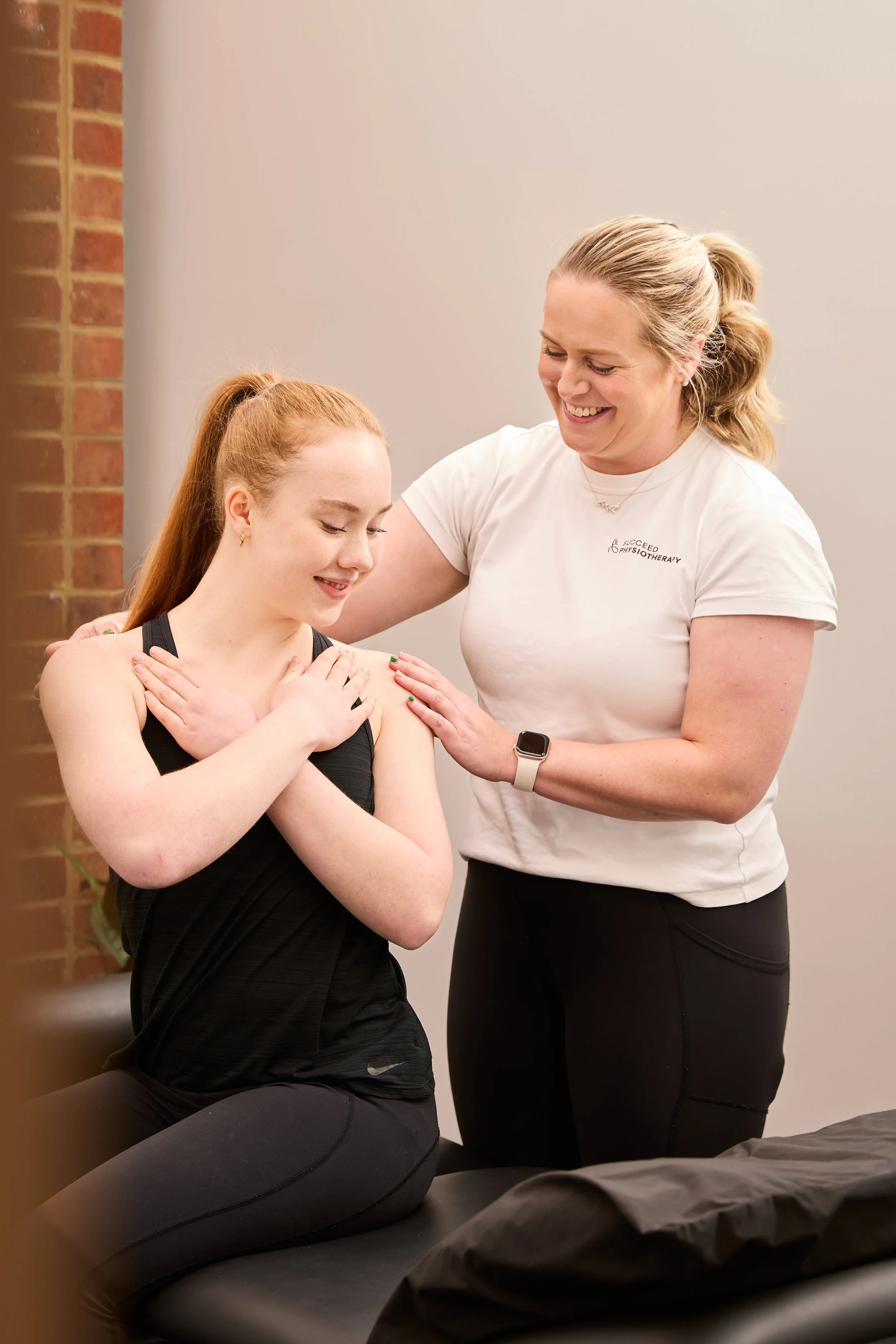 A physical therapist is assisting a young woman with shoulder exercises. The young woman is seated on an examination table with her arms crossed over her chest, and the therapist is smiling, placing her hands on the woman's shoulders. The scene takes place in a clinical setting.