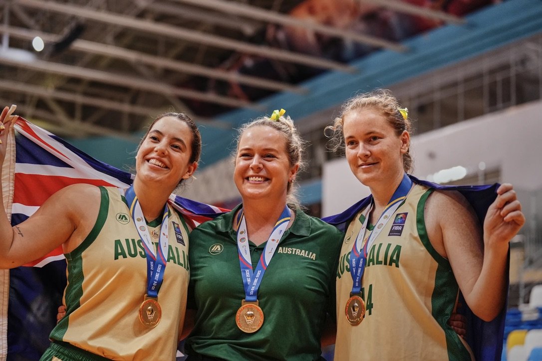 Three female athletes from Australia, wearing gold medals and sports uniforms, celebrating with their coach at a sports event.
