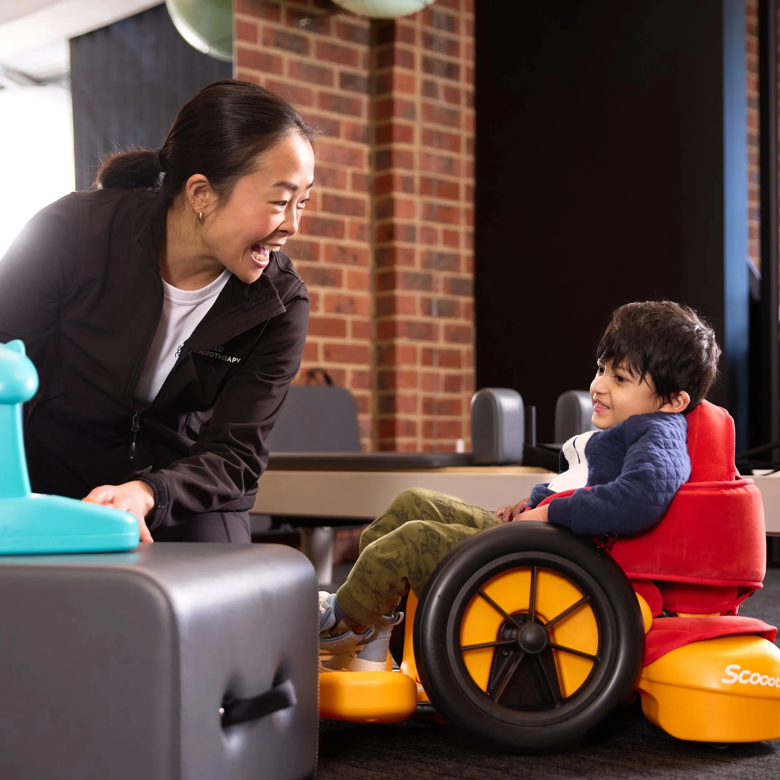 A woman and a young boy with a disability smiling at each other indoors with a brick wall in the background. The boy is in a red and yellow wheelchair.