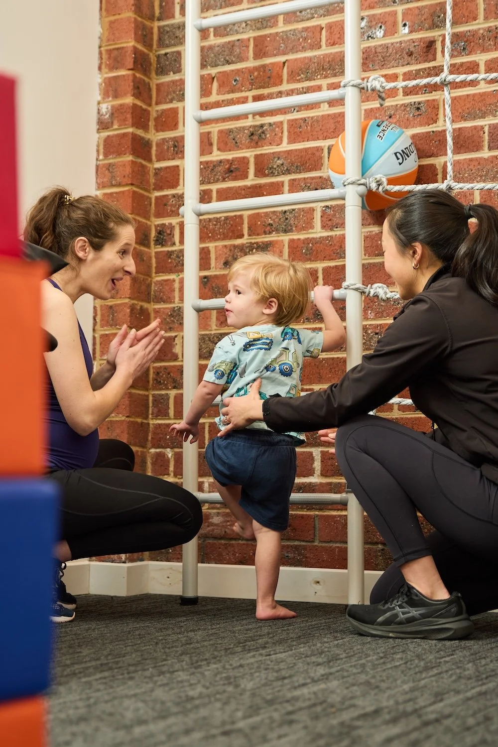 A young boy with red hair in shorts and a shirt with construction vehicle prints is climbing a gym ladder wall while two women, one on each side, help and encourage him in a gym with a brick wall background and a basketball attached to the wall.