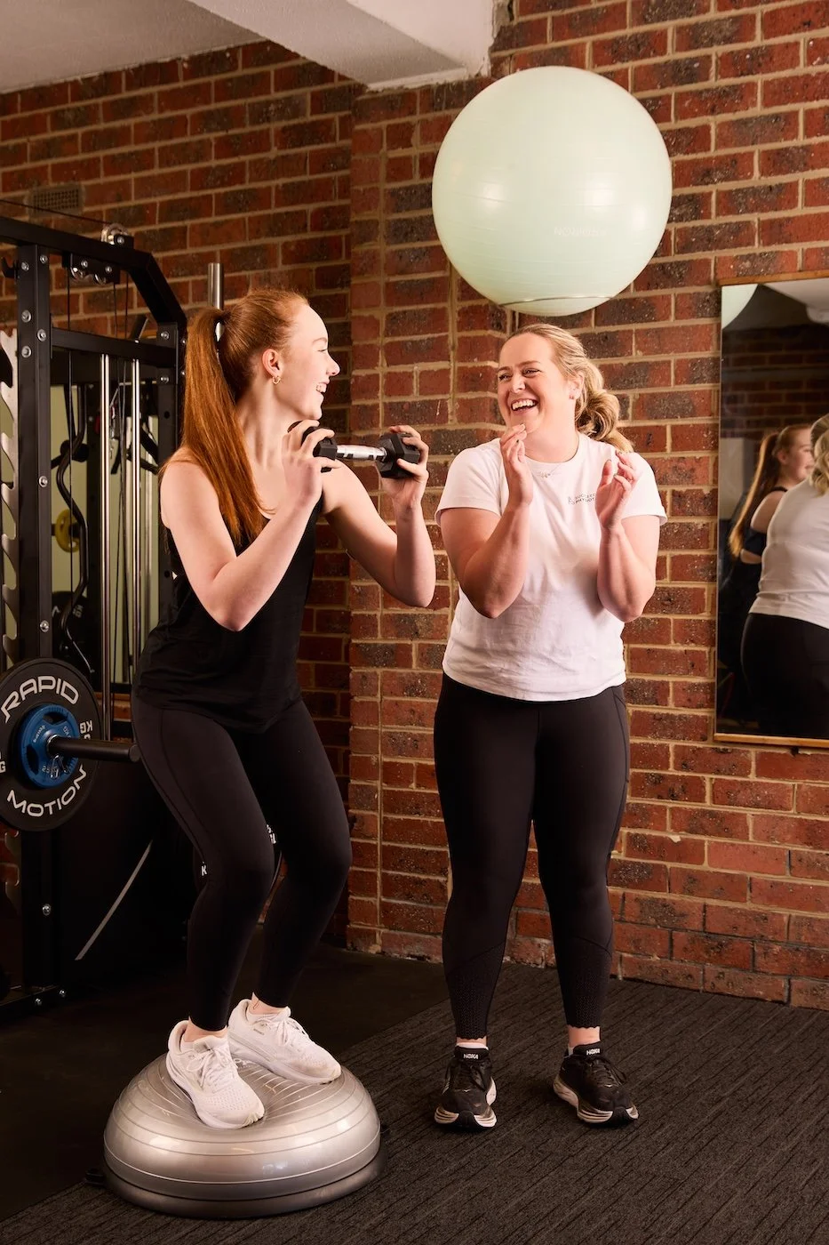 Two women laughing and smiling during a workout, one standing on a balance disk holding a dumbbell, the other clapping, in a gym with brick walls and mirrors.