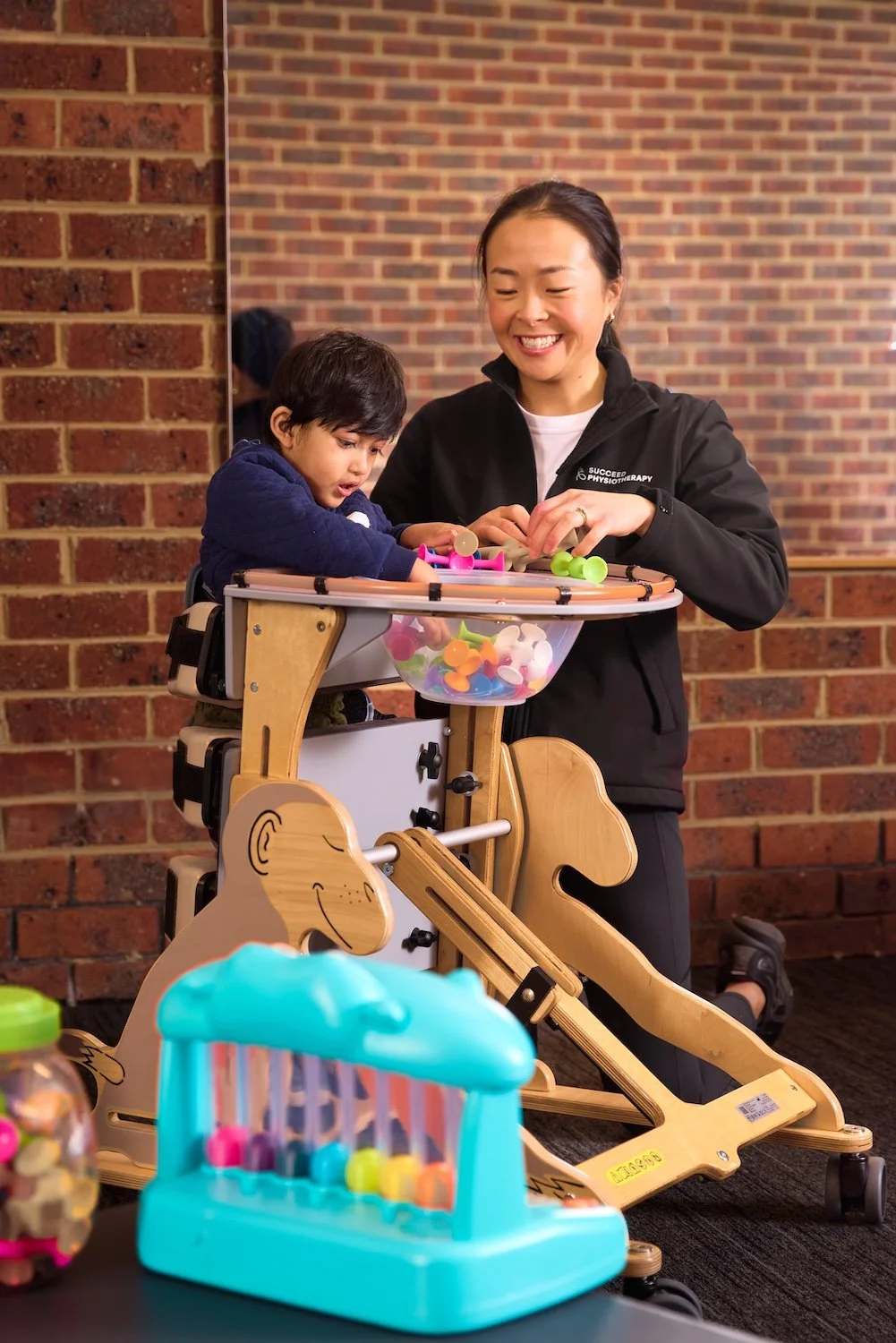 A woman and a young child smile at a table with colorful toys and a wooden playset in front of a brick wall.