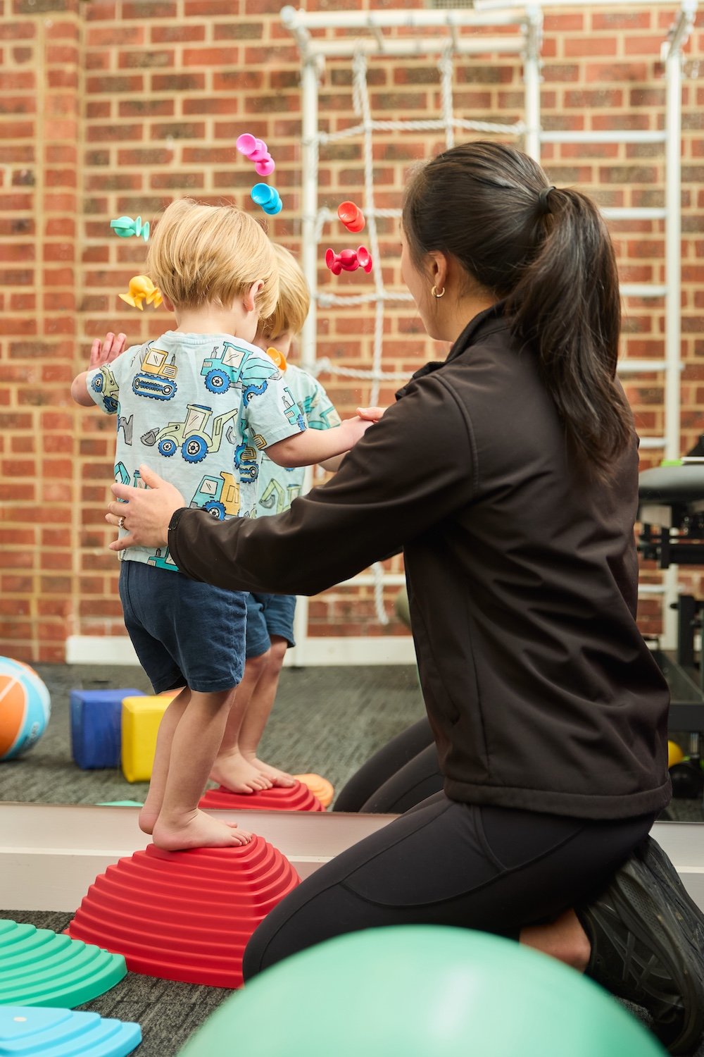 A woman kneeling and helping a young boy balance on a red, dome-shaped stepping stone in a therapy or gym setting. Two other children are in the background, and colorful therapy balls and blocks are on the floor, with a brick wall and climbing structure behind.