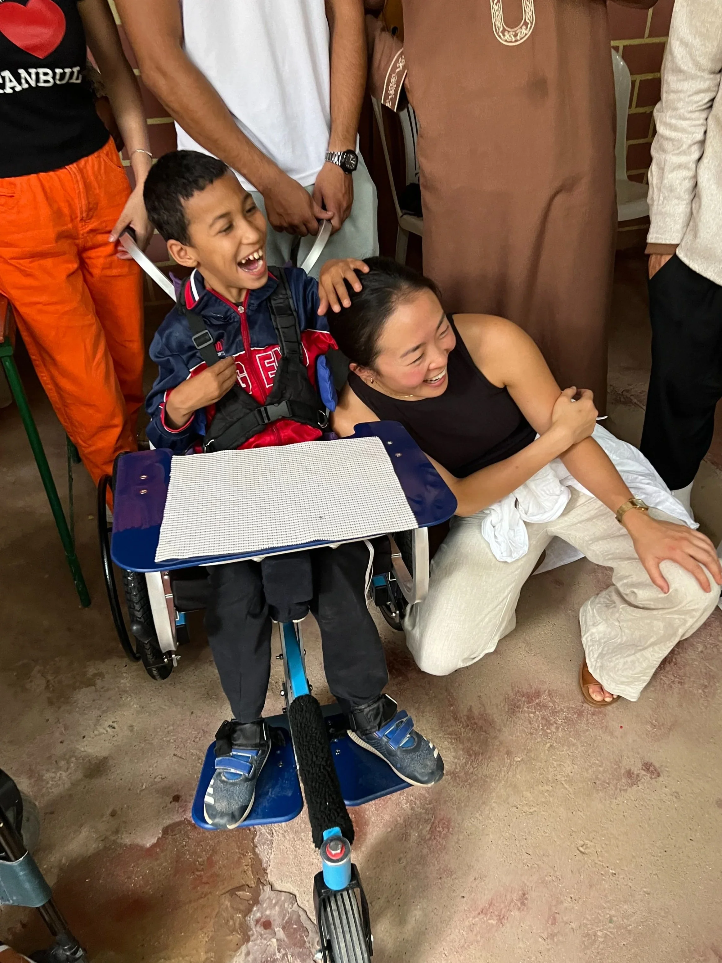 A young boy in a wheelchair and a woman are laughing together; the boy is touching the woman's head, and both are smiling. Several adults are standing around them, interacting and smiling.