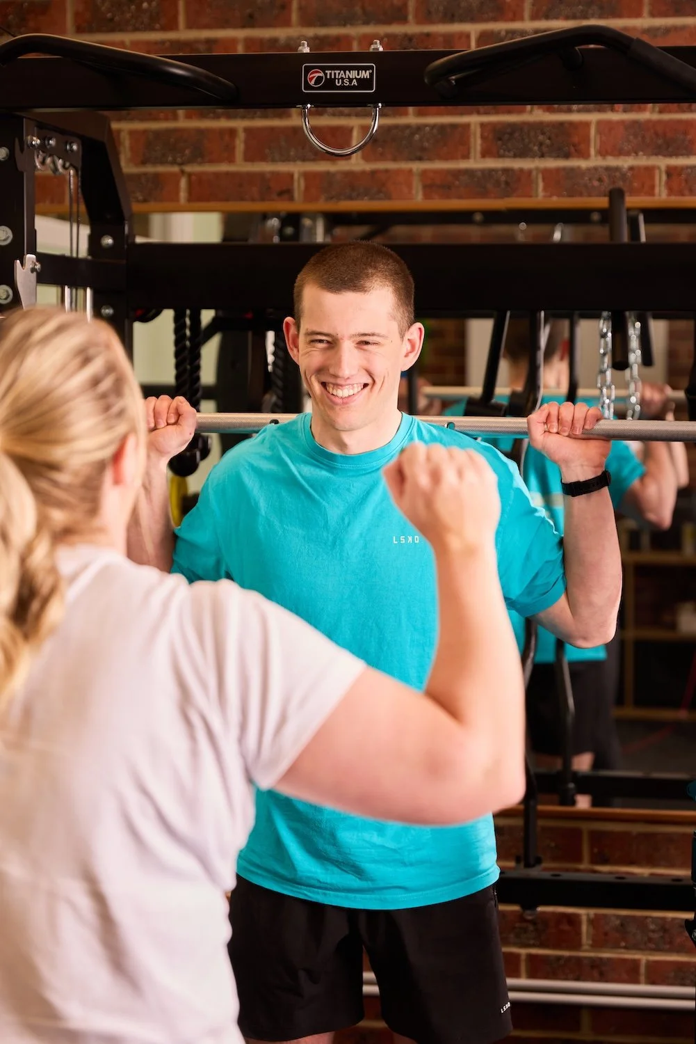 A young man smiling and chatting with a woman in a gym, holding a barbell on his shoulders.