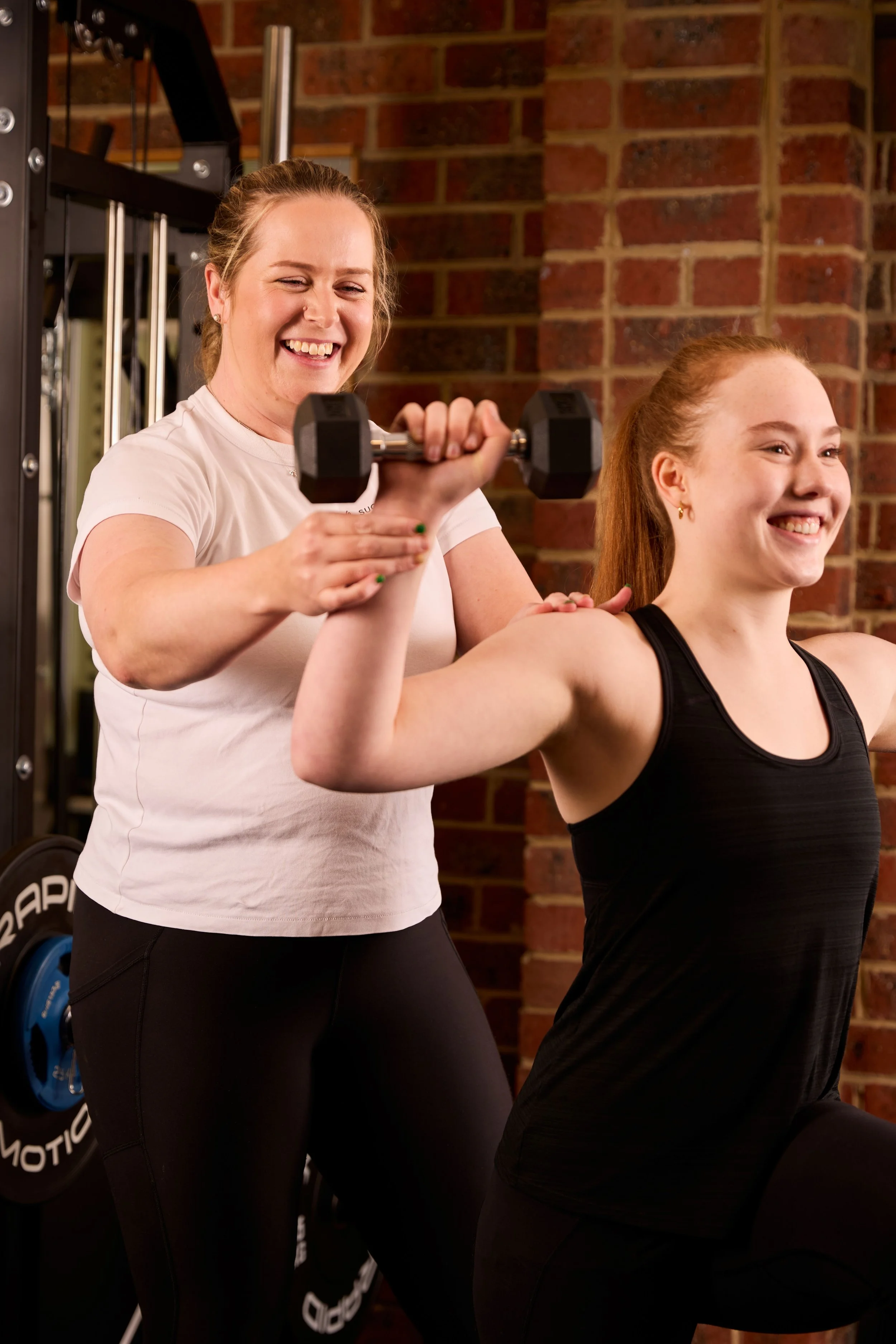 A woman is helping a young woman lift a dumbbell, smiling, in a gym with a brick wall background.