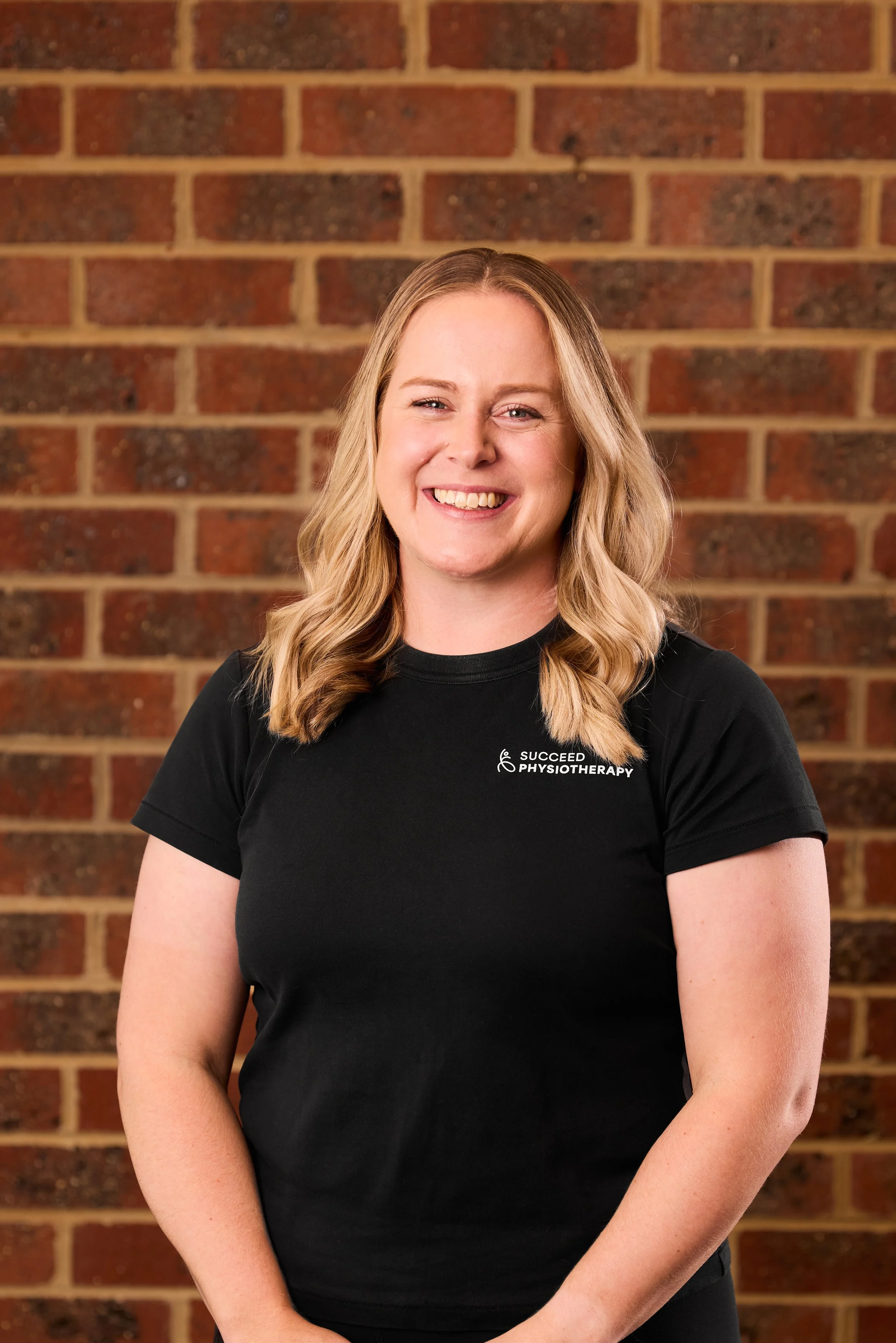 A woman with blonde hair smiling and wearing a black shirt that says 'SUCCEED PHYSIOTHERAPY' standing against a brick wall background.