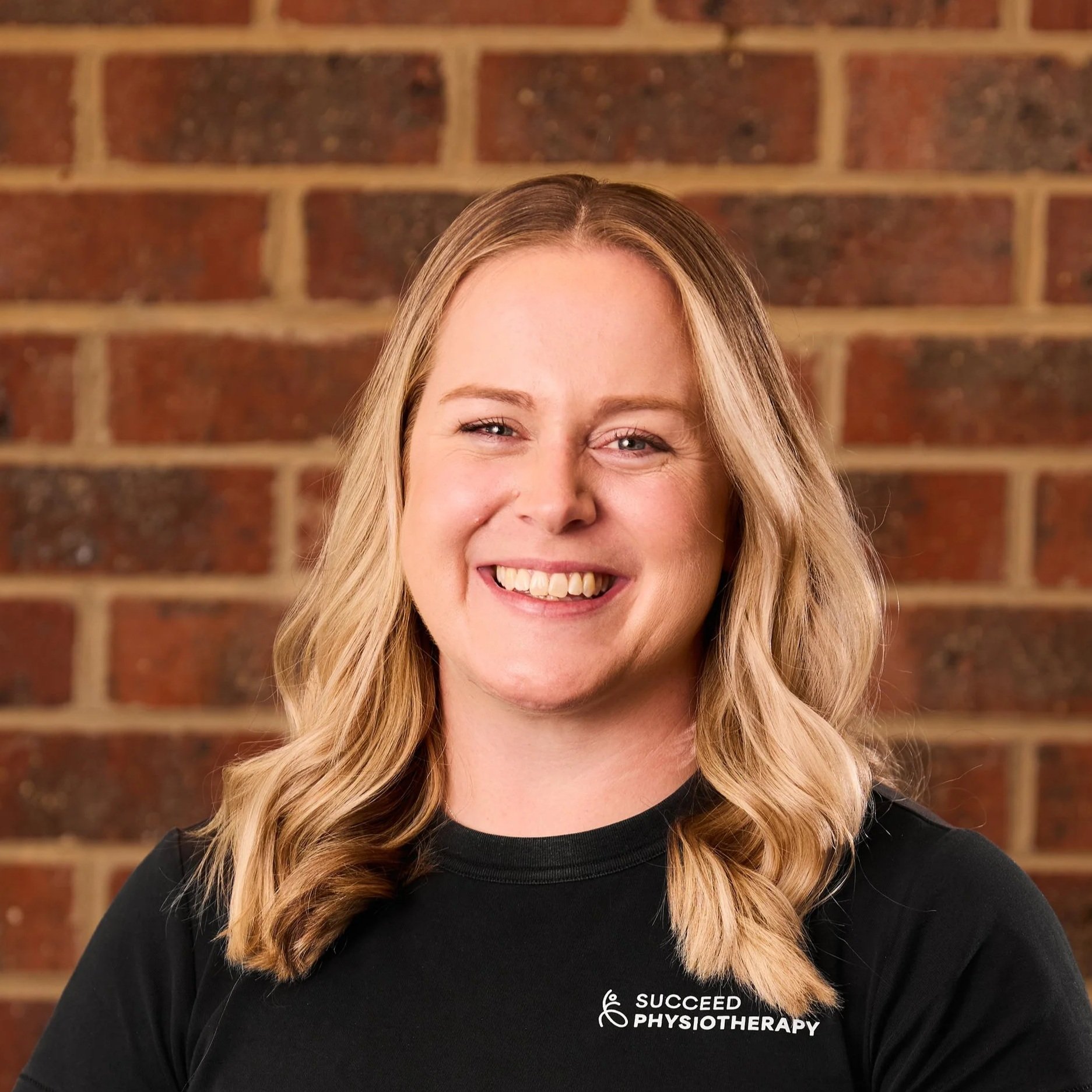 A smiling woman with shoulder-length blonde hair wearing a black T-shirt with the logo and text 'Succeed Physiotherapy' against a brick wall background.