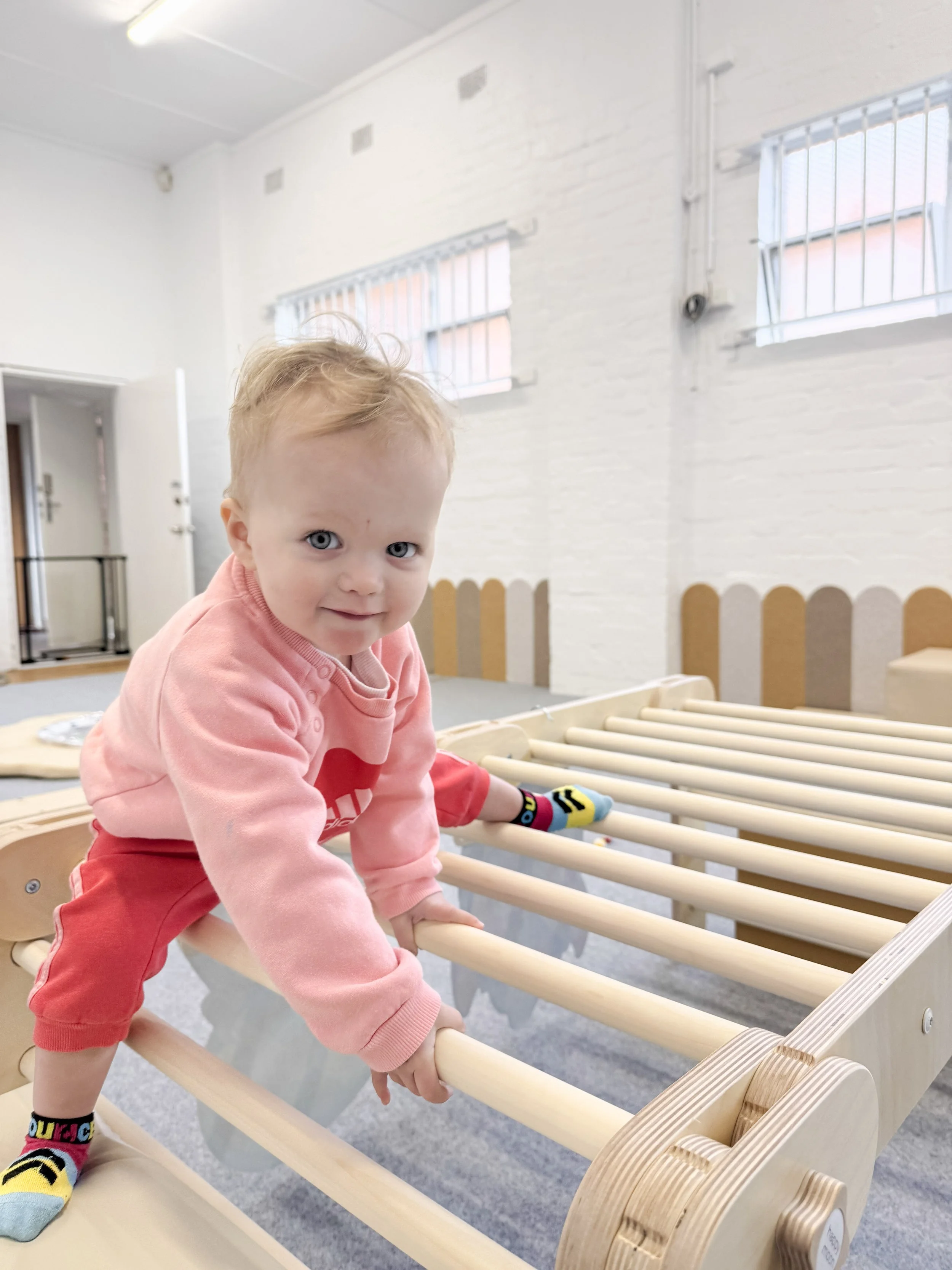 A young child with blonde hair and blue eyes, wearing a pink sweatshirt, red pants, and colorful socks, plays on a wooden slatted bed frame in a bright room with white brick walls and high windows.