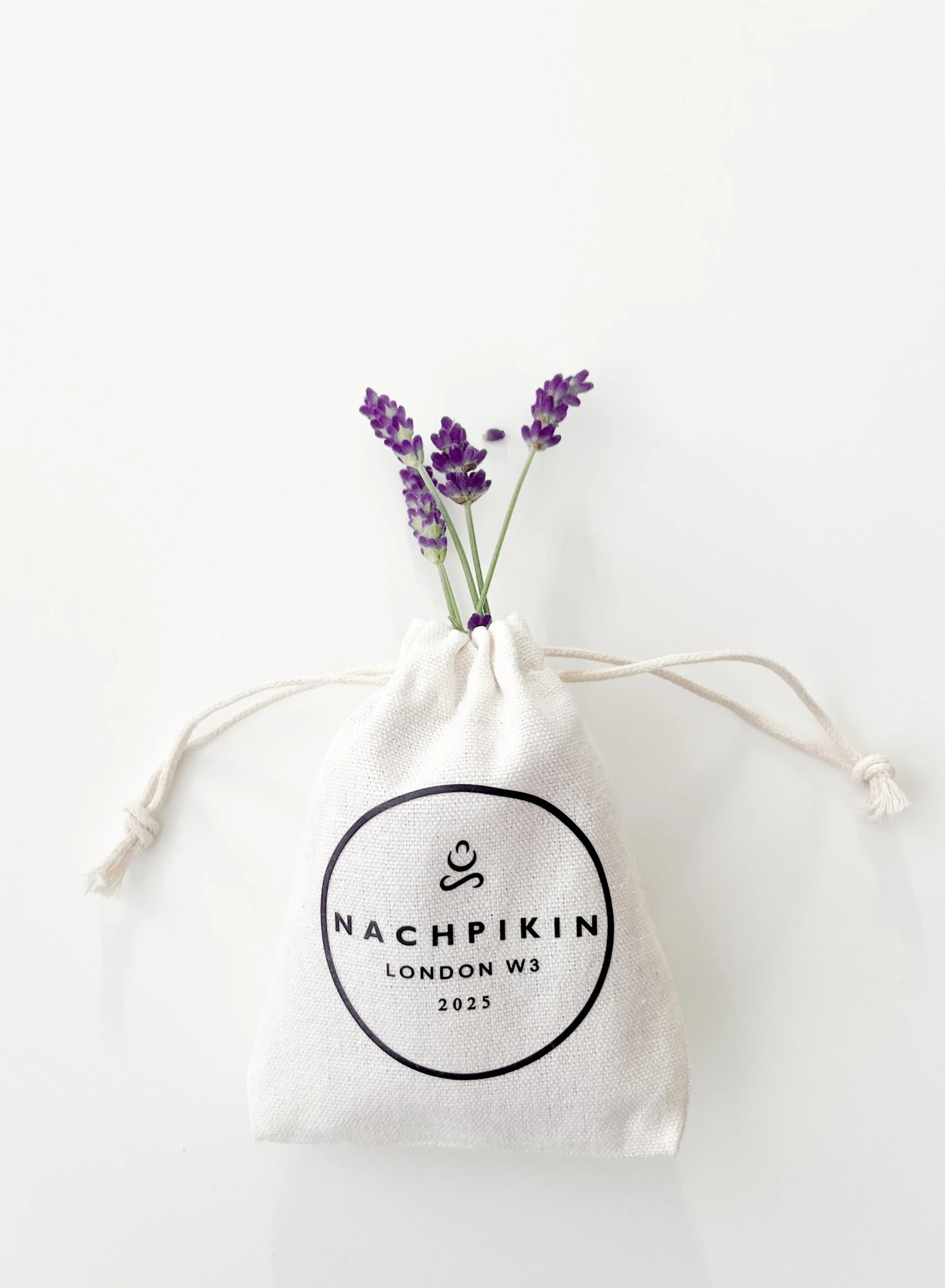 A small white drawstring pouch with the text 'NACHPIKIN LONDON W3 2025' printed on it, containing three sprigs of purple lavender flowers.
