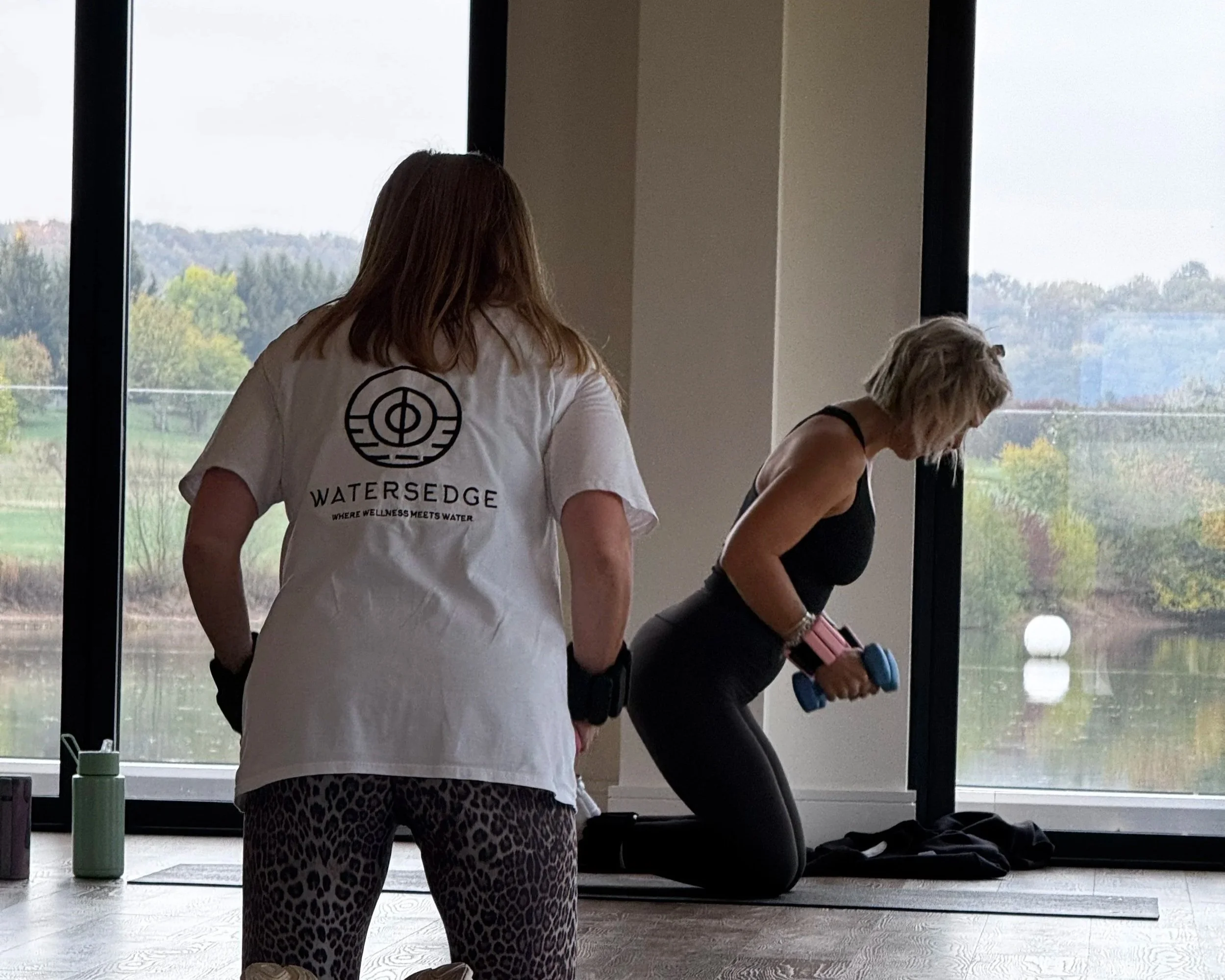 A woman in black workout clothes kneels on a yoga mat, holding pink and blue dumbbells, while a trainer in leopard-print leggings and a white t-shirt with 'WATERSEDGE' logo watches; large windows overlook a lake and trees.