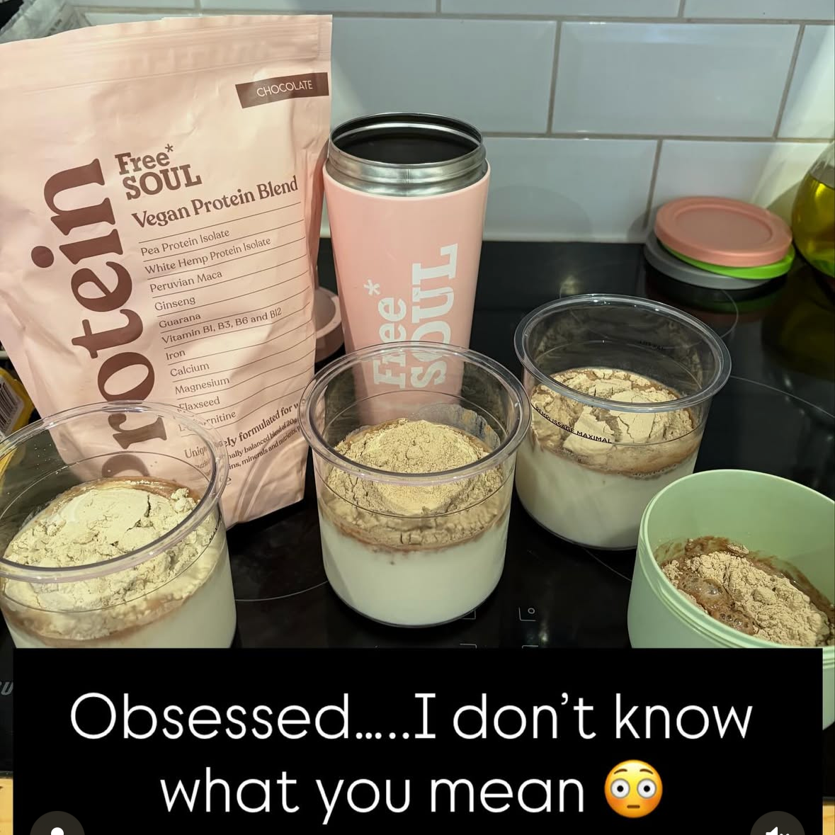 Three cups with layered protein smoothies topped with powder, a bag of plant-based protein powder labeled Free Soul Vegan Protein Blend, a pink tumbler with the message "Feed * Soul," and a small green container with powder on a kitchen counter with a tiled wall in the background.