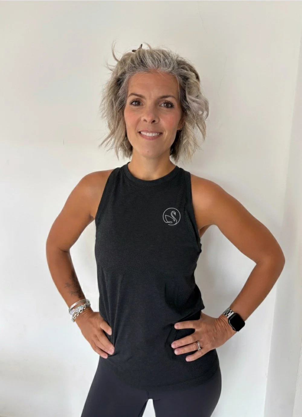 Woman with gray hair smiling, wearing a black sleeveless workout top and black leggings, standing with hands on hips against a plain white wall.