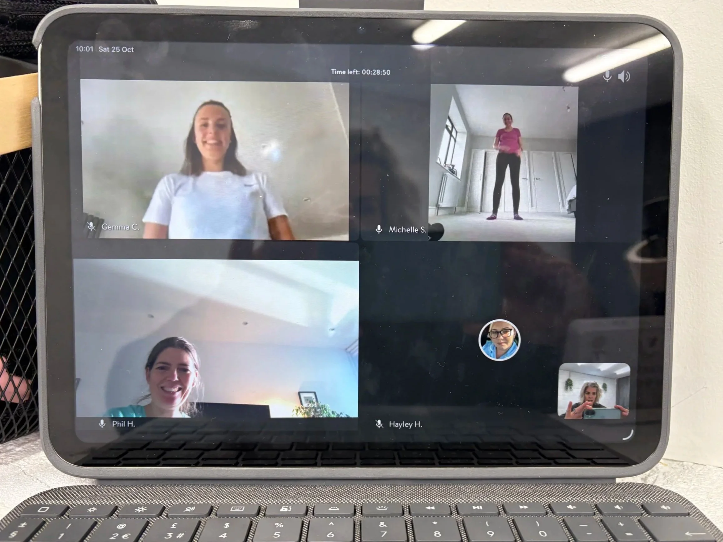 A tablet on a desk showing a virtual meeting with five women in separate video call windows. Four women, including one in a white shirt and another in a teal shirt, are in different indoor spaces, some smiling. The fifth participant appears as a small circular icon of a woman with glasses.